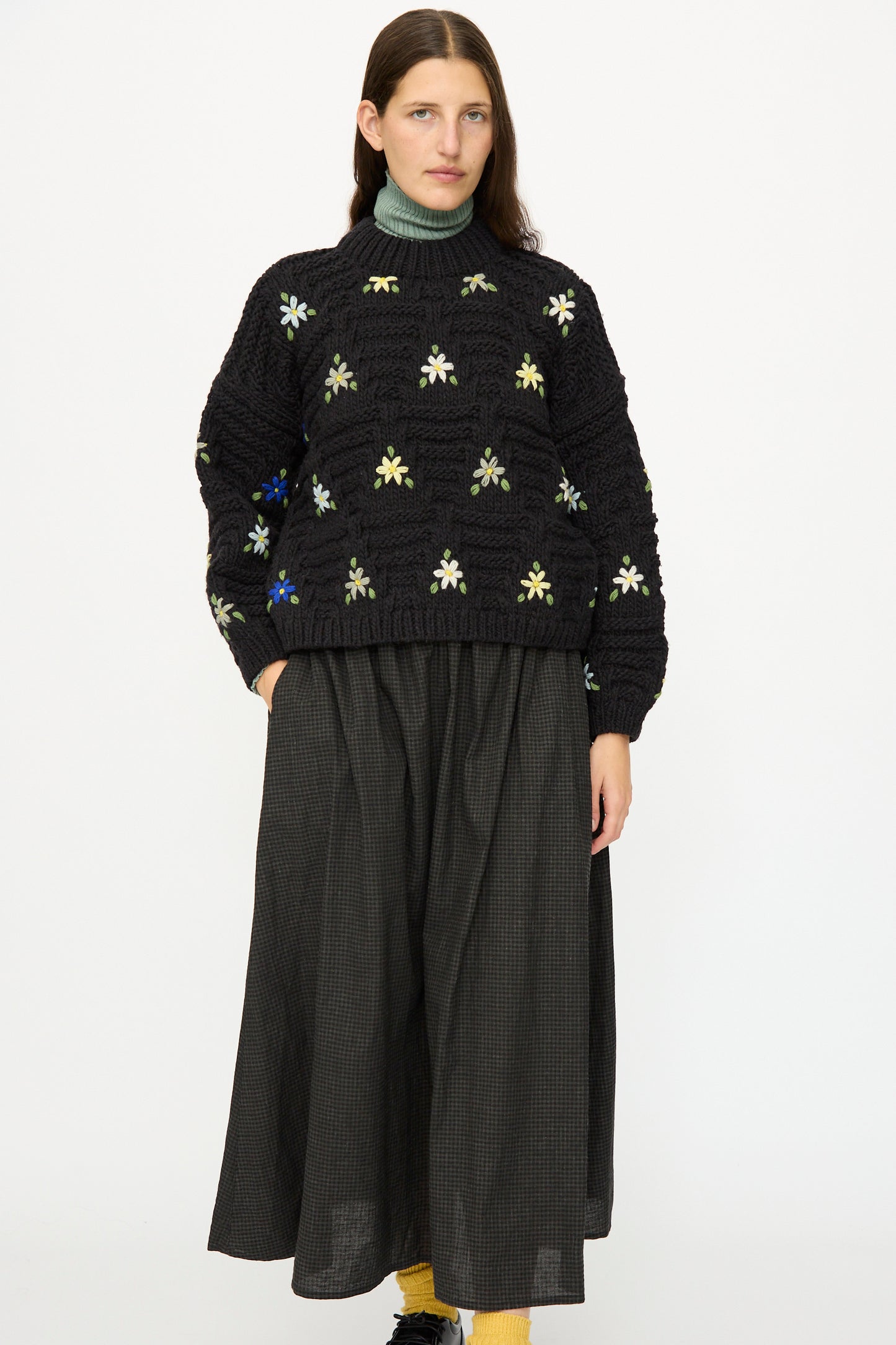 A woman poses against a white background wearing the Ichi Antiquités Wool Hand-knit Pullover with Flowers in Black, styled over a green turtleneck and a long black skirt.