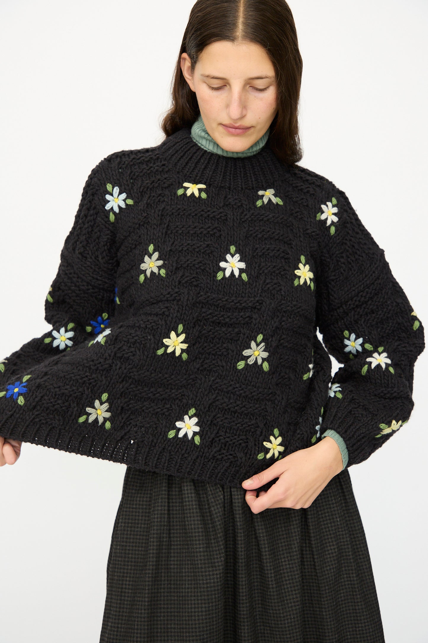 A woman wears the Ichi Antiquités Wool Hand-knit Pullover with Flowers in Black, paired with a dark checkered skirt, standing against a plain background.