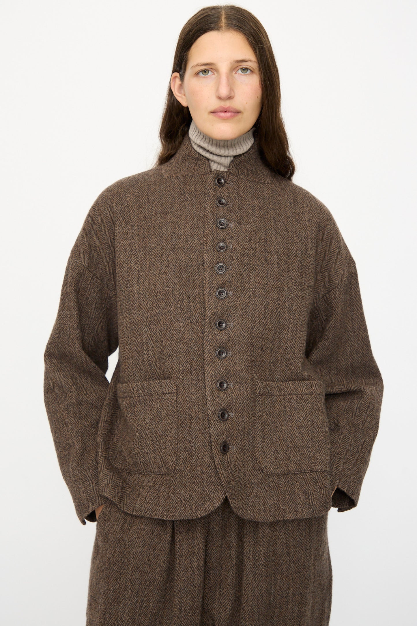 A person with long brown hair wears the Ichi Antiquités Wool Herringbone Jacket in Brown over a beige turtleneck, paired with matching pants, standing against a plain white background.