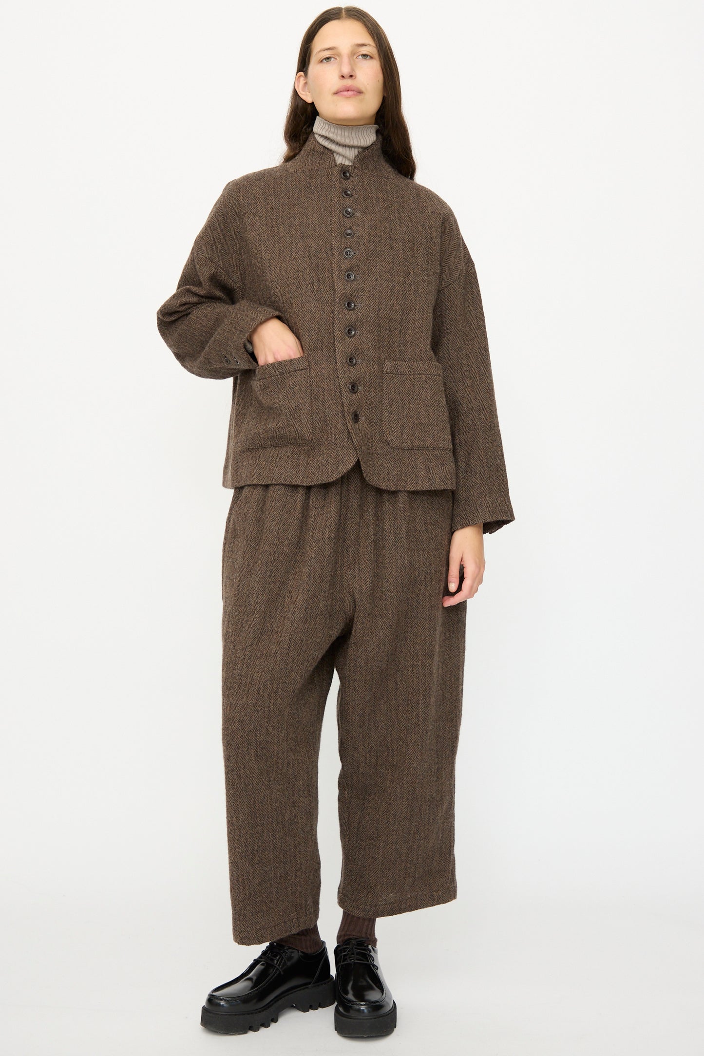 A person stands against a white background wearing the Ichi Antiquités Wool Herringbone Jacket in Brown, paired with matching wide-leg trousers, a beige turtleneck, and black shoes.