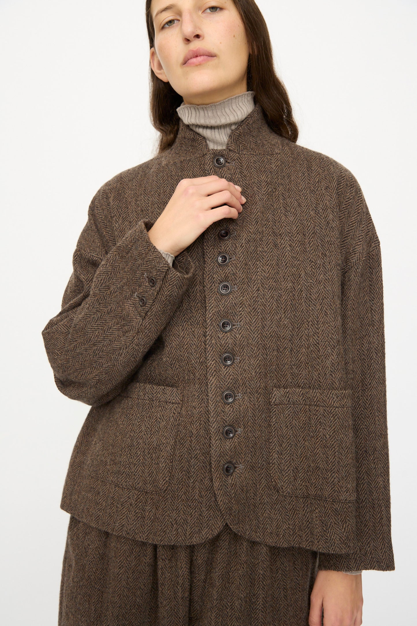A person wearing the Ichi Antiquités Wool Herringbone Jacket in Brown over a beige turtleneck, with one hand touching the collar.