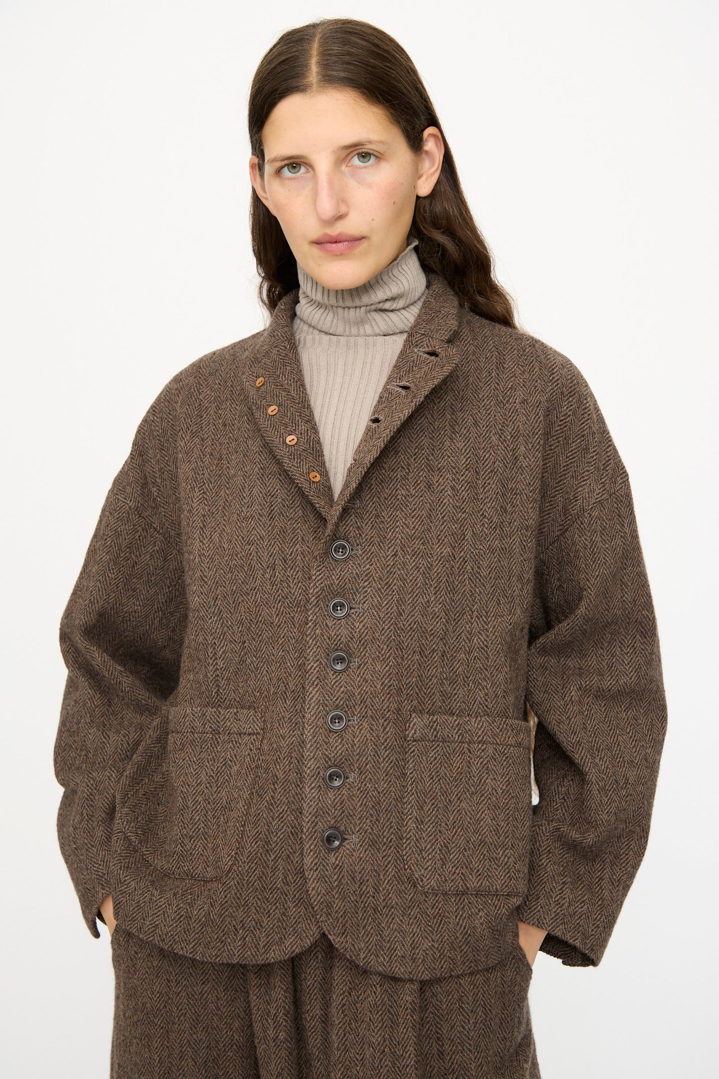 A person with long brown hair models the Ichi Antiquités Wool Herringbone Jacket in Brown, layered over a taupe turtleneck, against a plain white background.