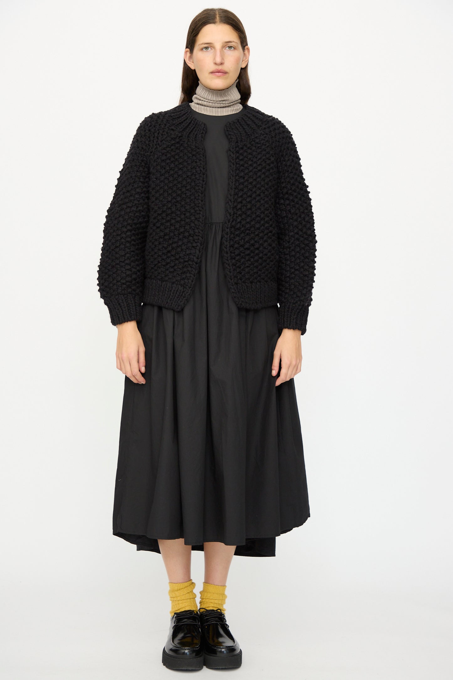 A person wears the Ichi Antiquités Wool Peru Hand-knit Cardigan in Black over a black midi dress, paired with yellow socks and black shoes, standing against a plain white background.
