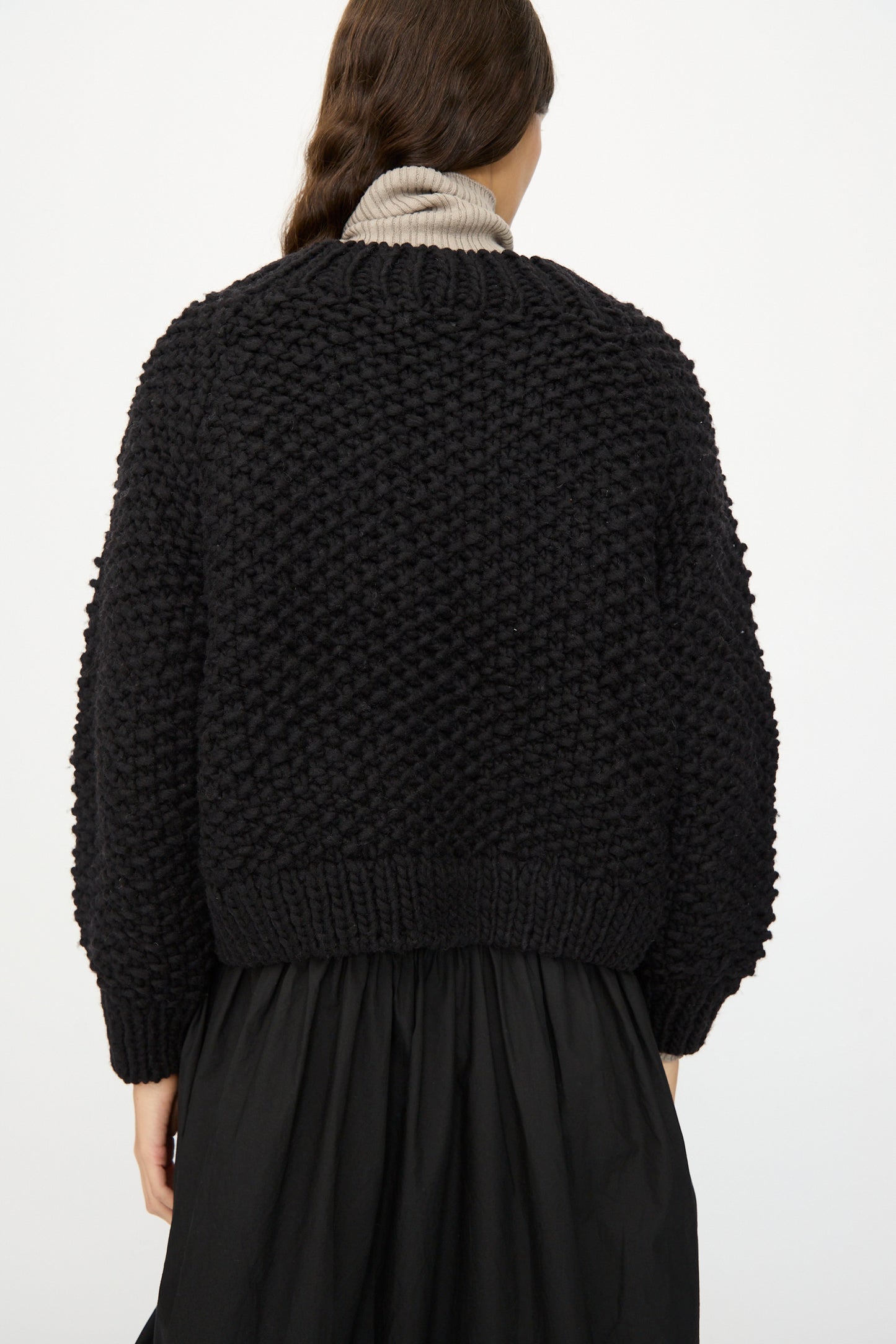A person with long brown hair, seen from behind, wears the Ichi Antiquités Wool Peru Hand-knit Cardigan in Black over a beige turtleneck and black skirt against a plain background.