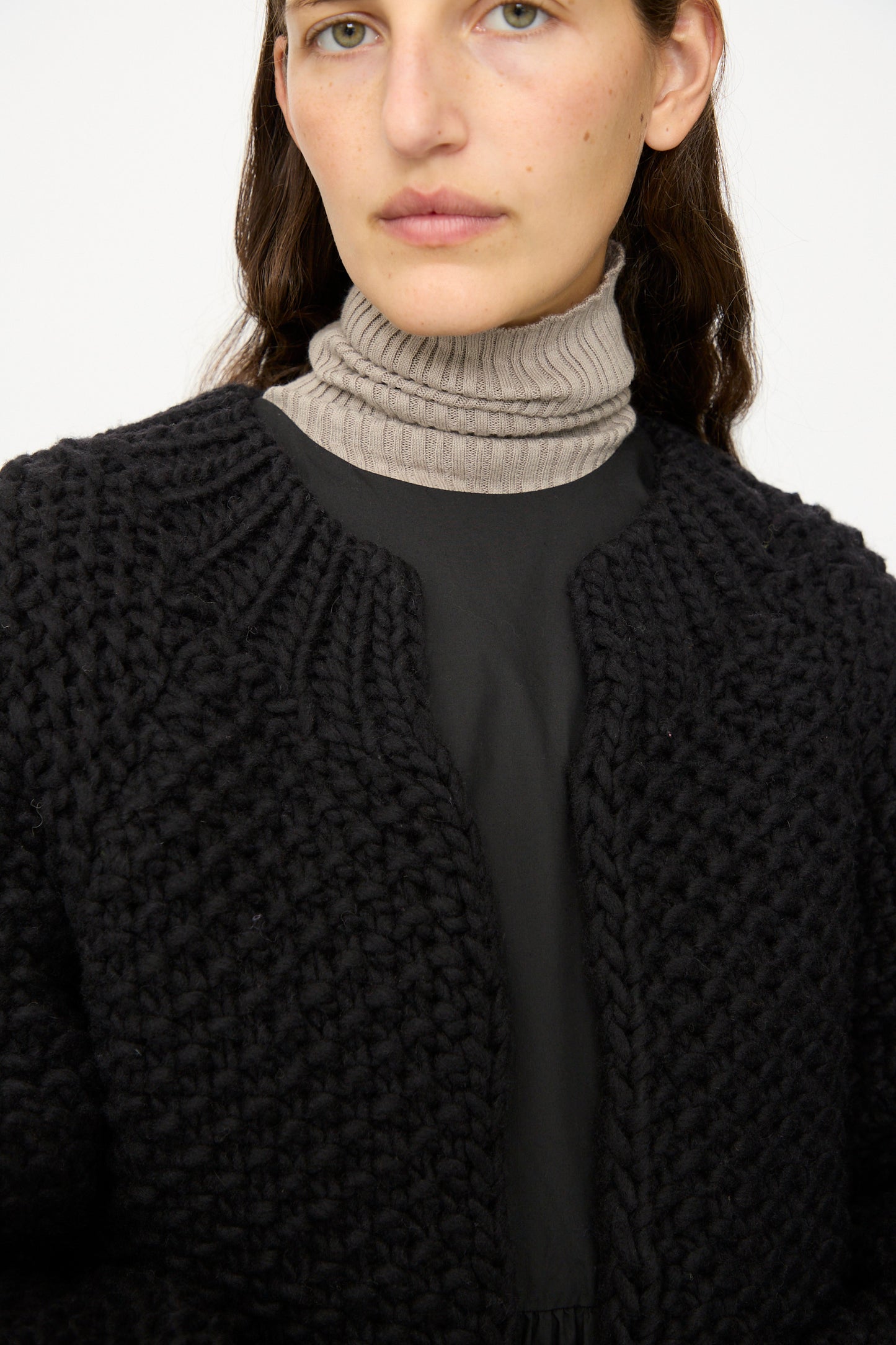 A person wears the Ichi Antiquités Wool Peru Hand-knit Cardigan in Black over a black top and beige ribbed turtleneck, facing forward with a neutral expression.