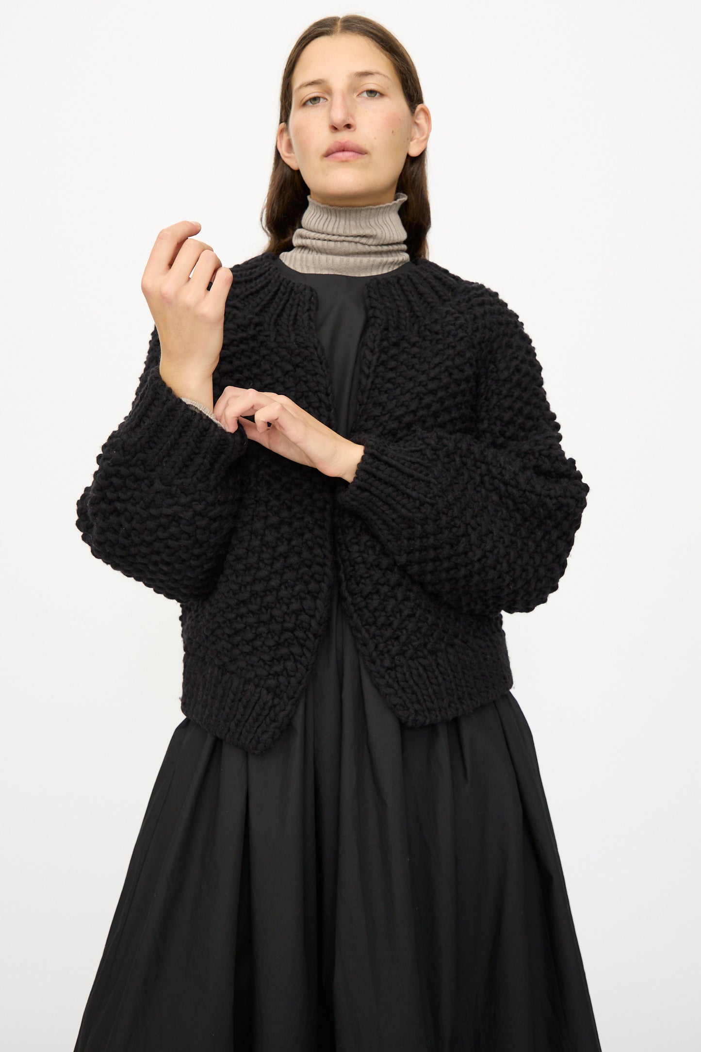 A model wears the Ichi Antiquités Wool Peru Hand-knit Cardigan in Black over a gray turtleneck and black dress, standing against a plain white background.