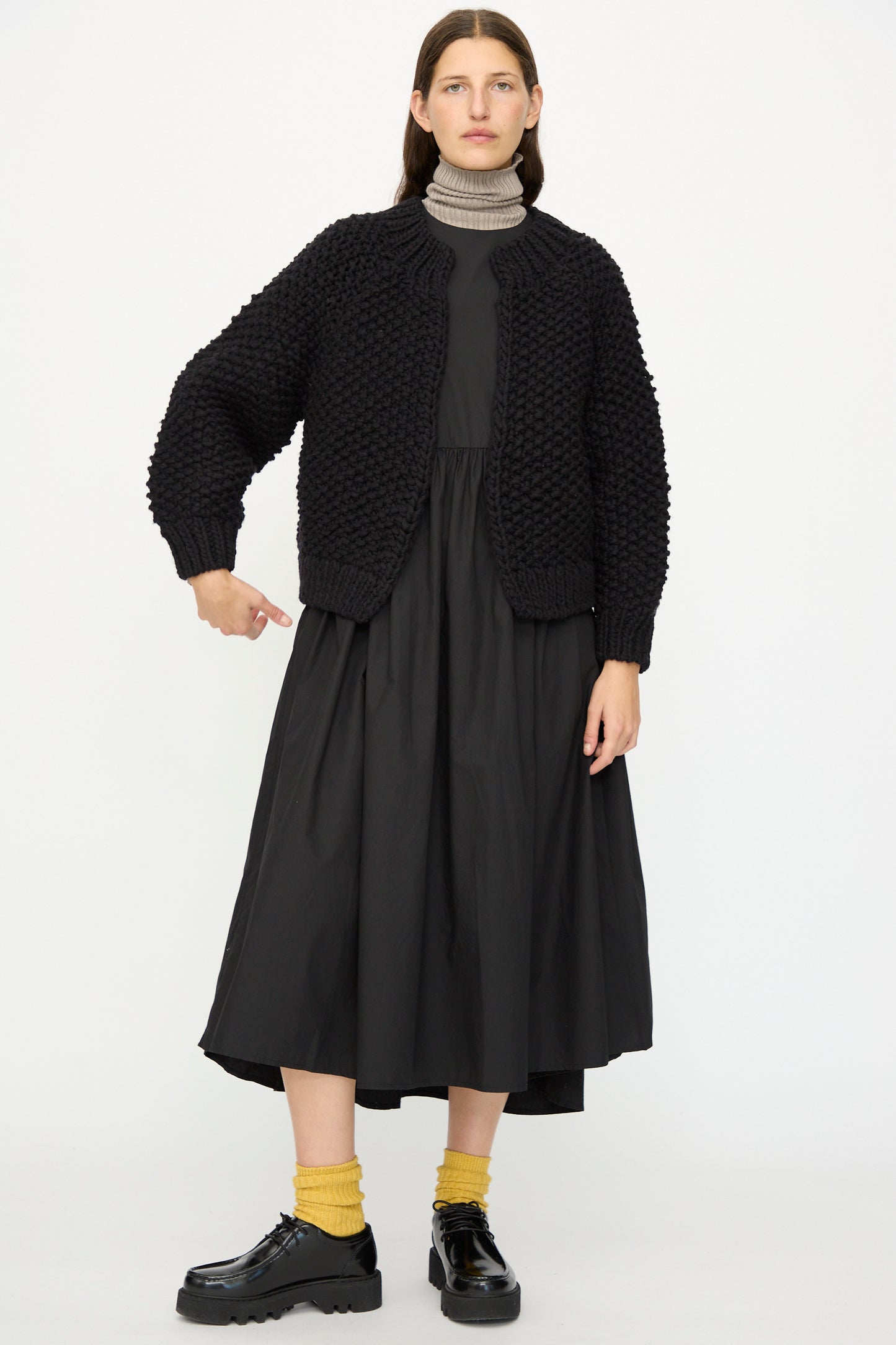 A woman stands against a white background wearing the Ichi Antiquités Wool Peru Hand-knit Cardigan in Black, a long black dress, yellow socks, and black platform shoes, pointing downward with her right hand.