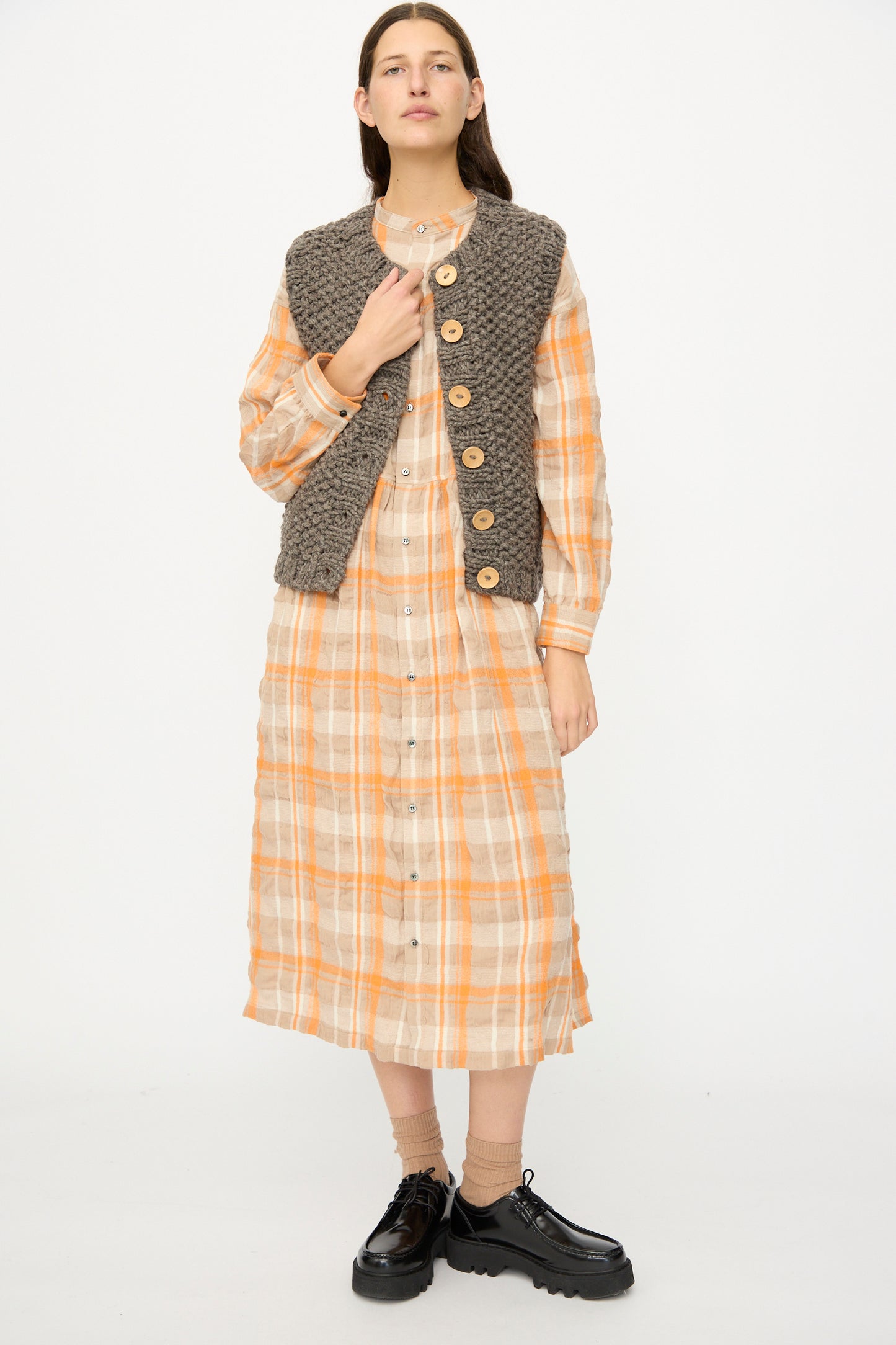 A person stands against a plain white background wearing a long orange and beige plaid dress with buttons, the Wool Peru Hand-knit Vest in Gray by Ichi Antiquités, beige socks, and black platform shoes.