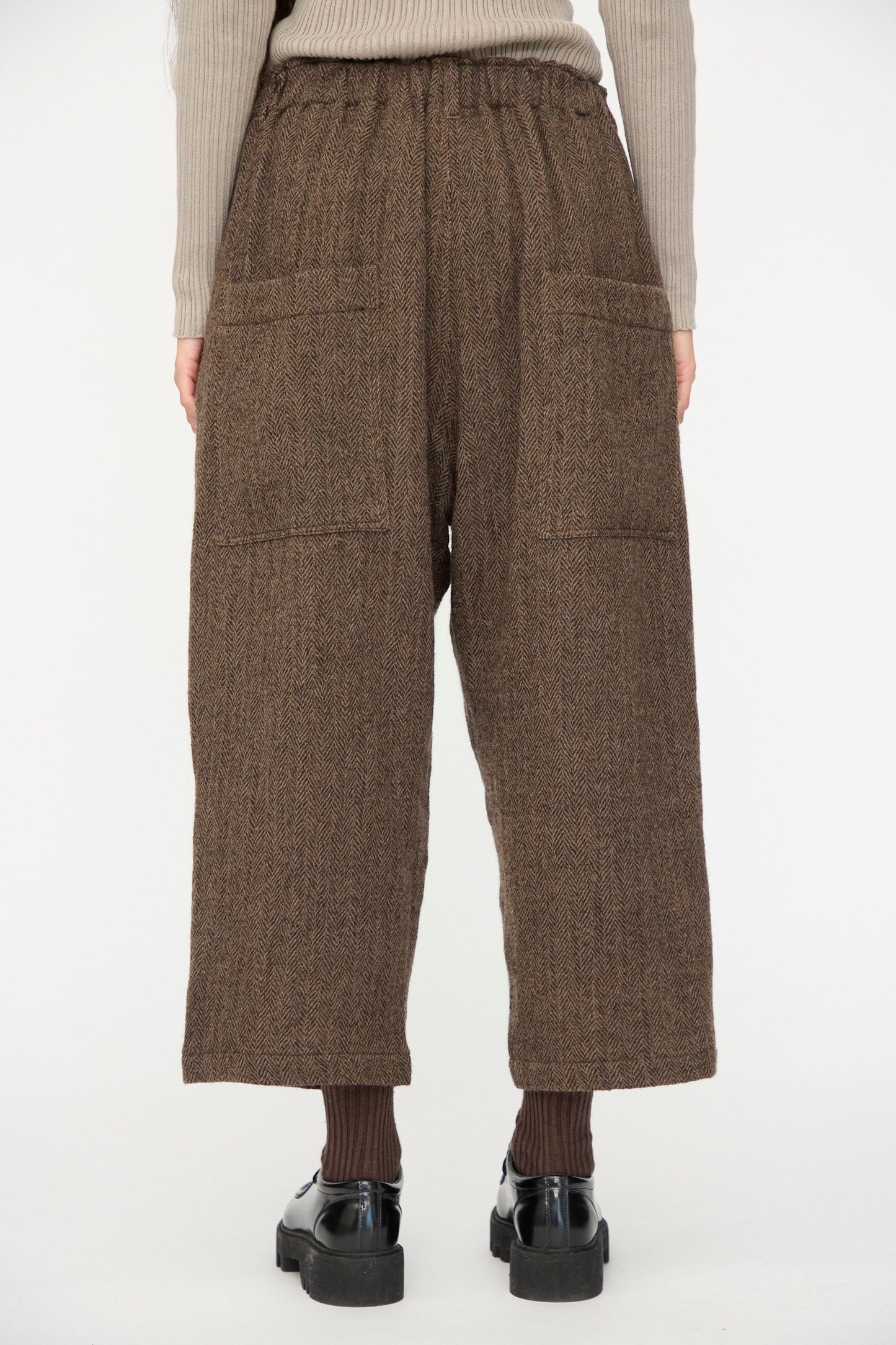 Model wears Ichi Antiquités Wool Herringbone Pants in Brown, featuring two large back pockets, paired with a ribbed beige top, brown socks, and black shoes; shown from the back against a plain background.