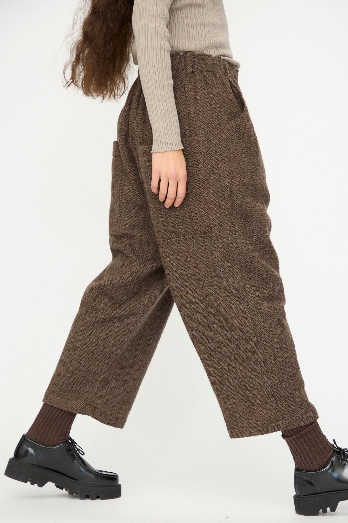 A person wearing beige ribbed sweater, brown socks, black loafers, and Ichi Antiquités Wool Herringbone Pants in Brown walks against a plain white background.