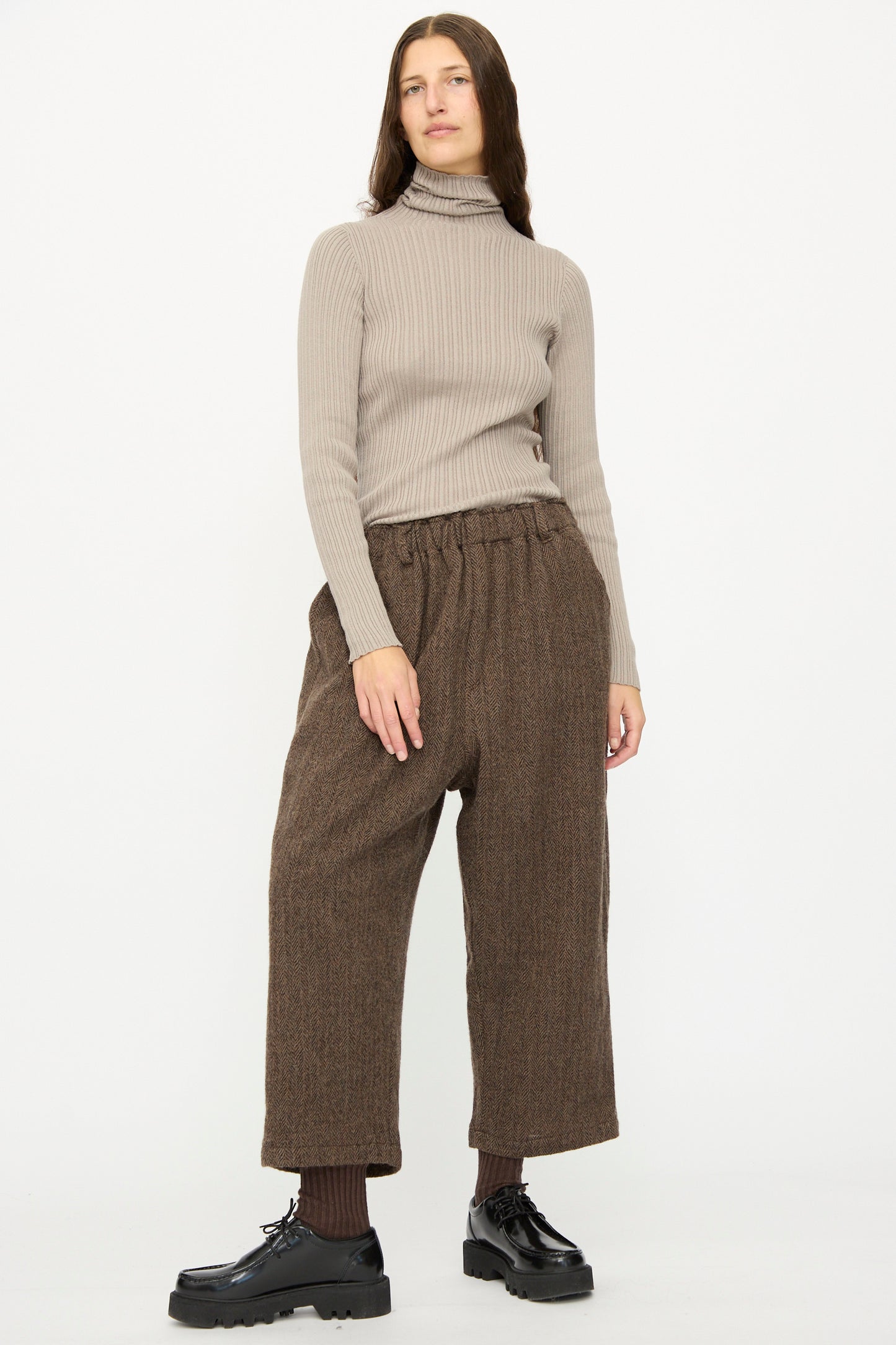 A person stands against a white background wearing a beige ribbed turtleneck, dark socks, black shoes, and Ichi Antiquités Wool Herringbone Pants in Brown.