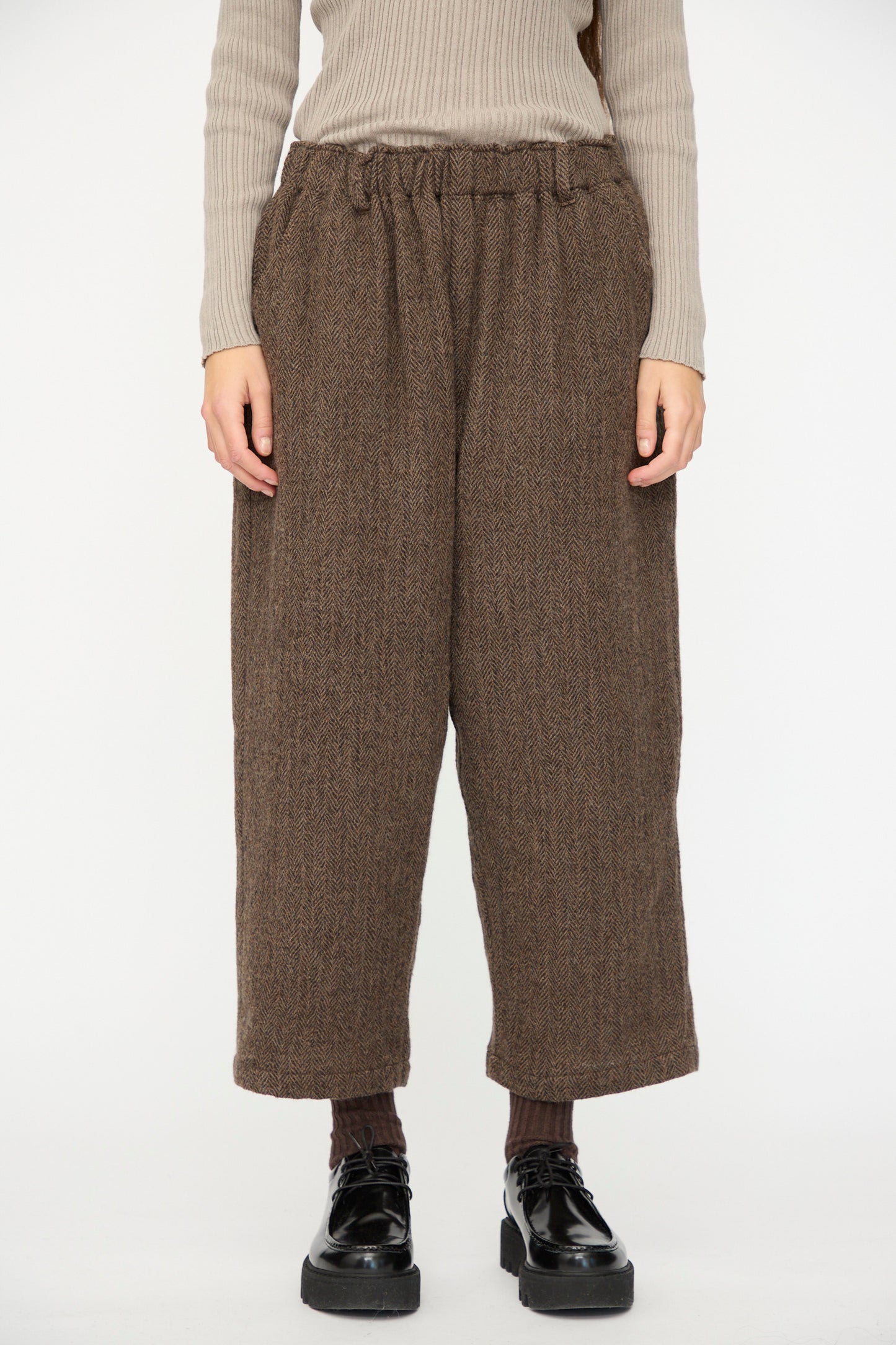 Person wearing Ichi Antiquités Wool Herringbone Pants in Brown, a ribbed beige long-sleeve top, dark socks, and black platform shoes, standing against a plain white background.