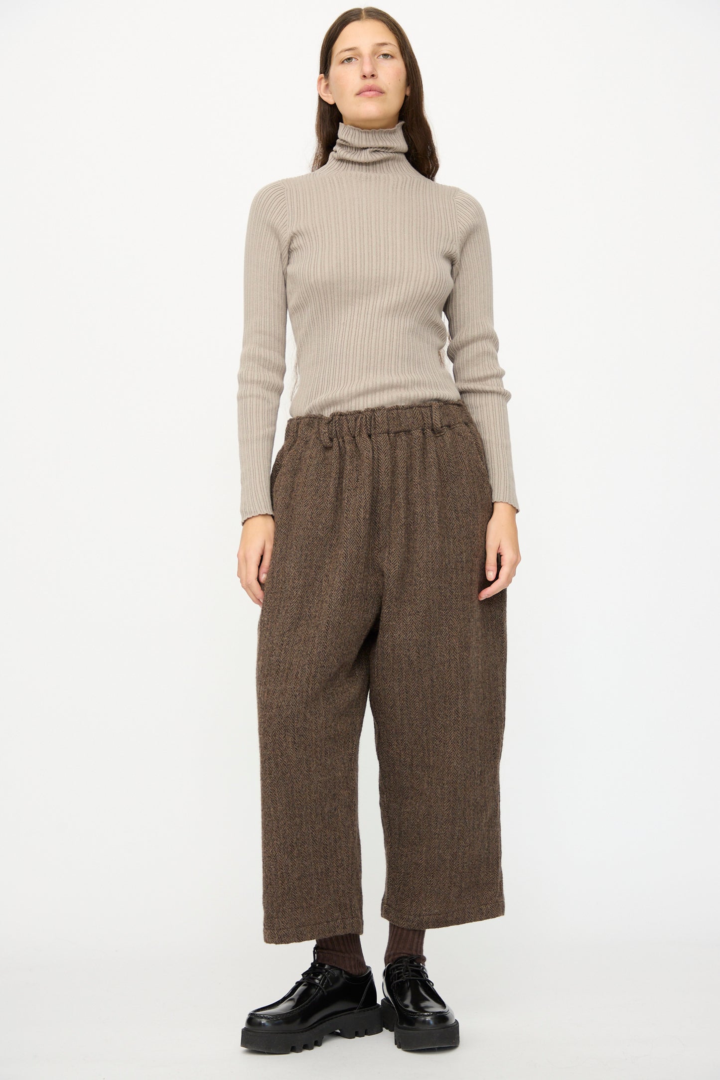 A person stands against a plain white background wearing Wool Herringbone Pants in Brown by Ichi Antiquités, paired with a beige ribbed turtleneck sweater, dark socks, and black shoes.