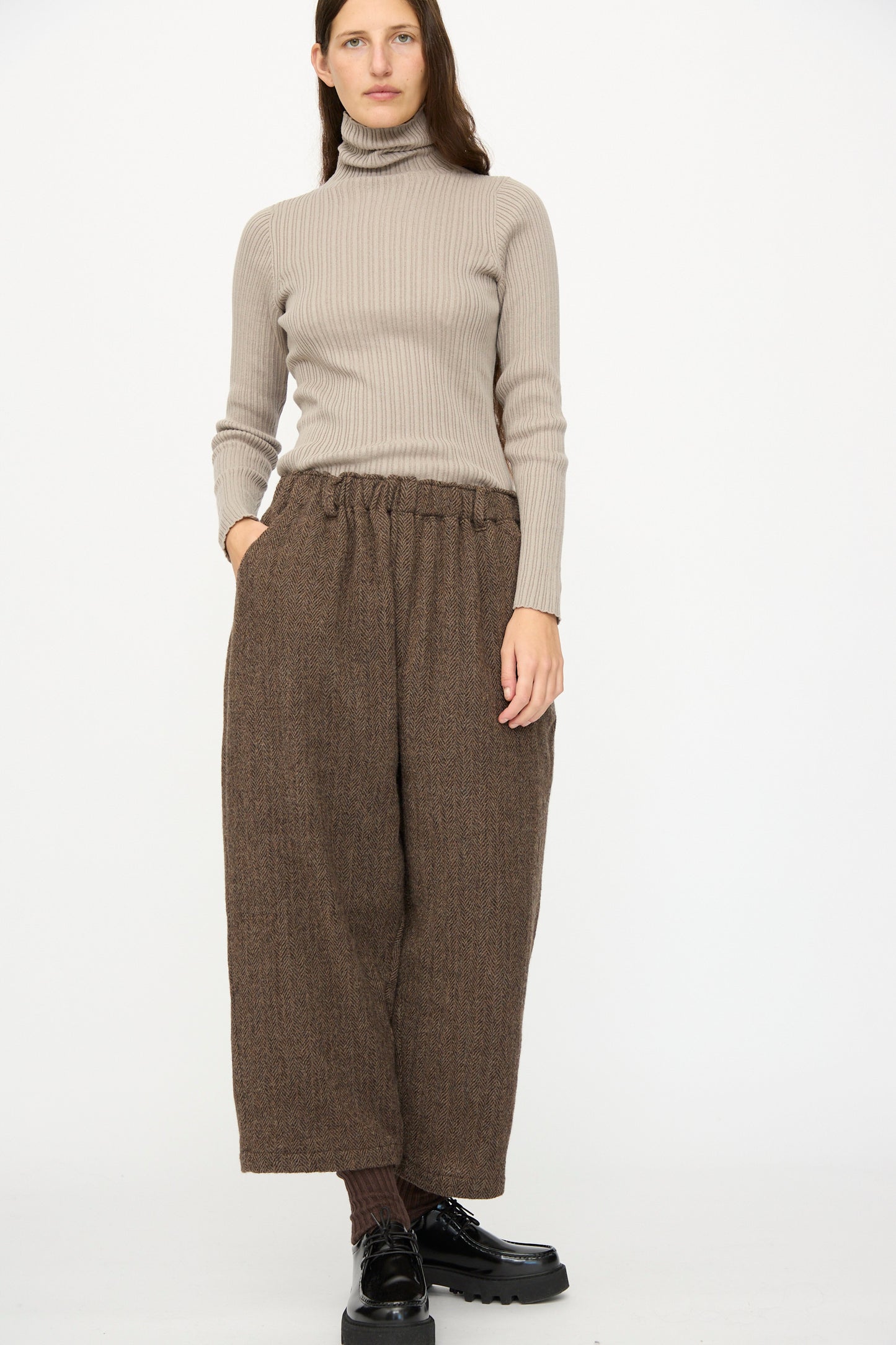 A person stands with one hand in their pocket, wearing a beige ribbed turtleneck, Ichi Antiquités Wool Herringbone Pants in Brown, brown socks, and black shoes against a plain white background.