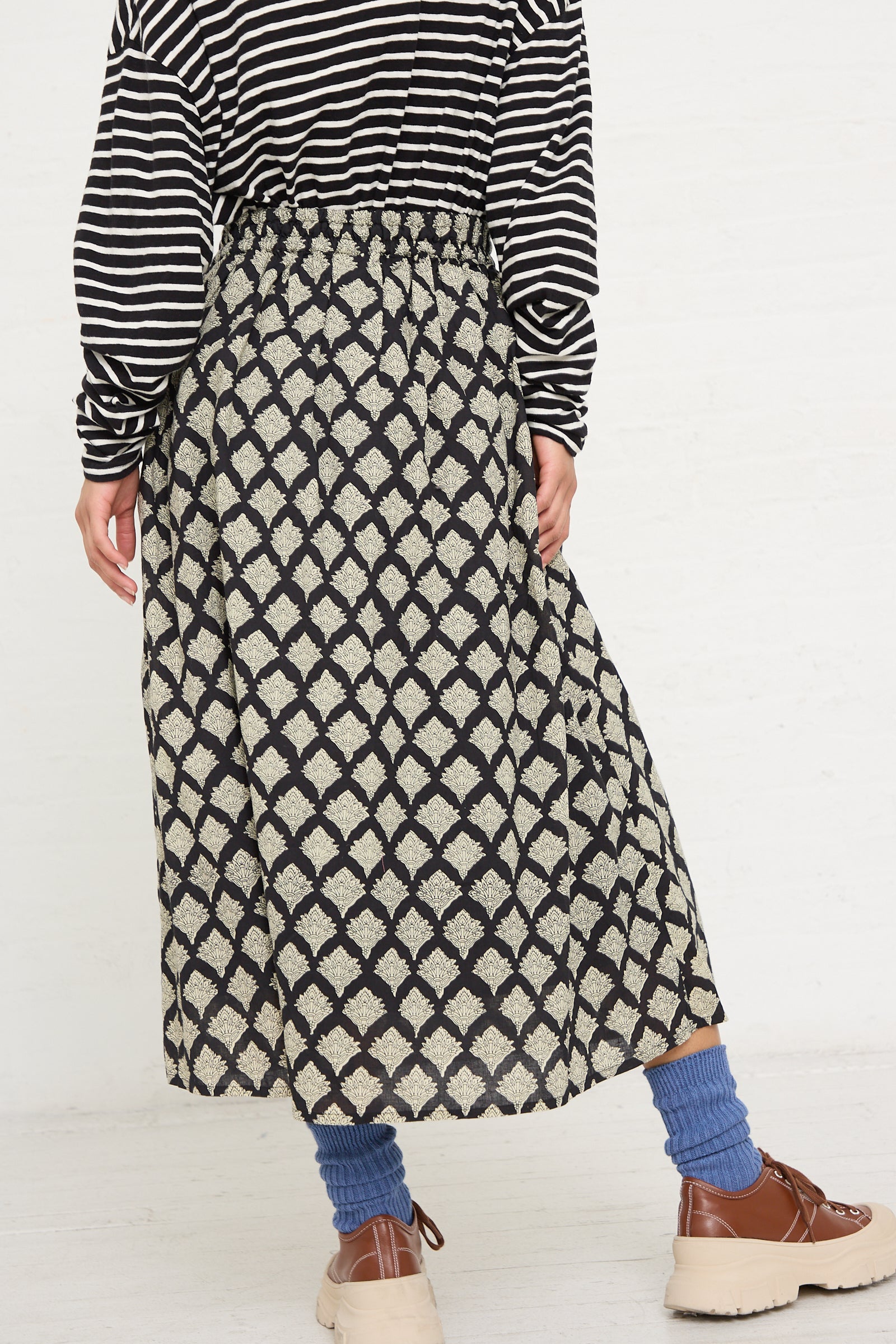 A person wearing a black and white striped top, an Ichi Antiquités Woven Cotton Indian Block Print Skirt in Black, blue socks, and brown shoes against a white background.