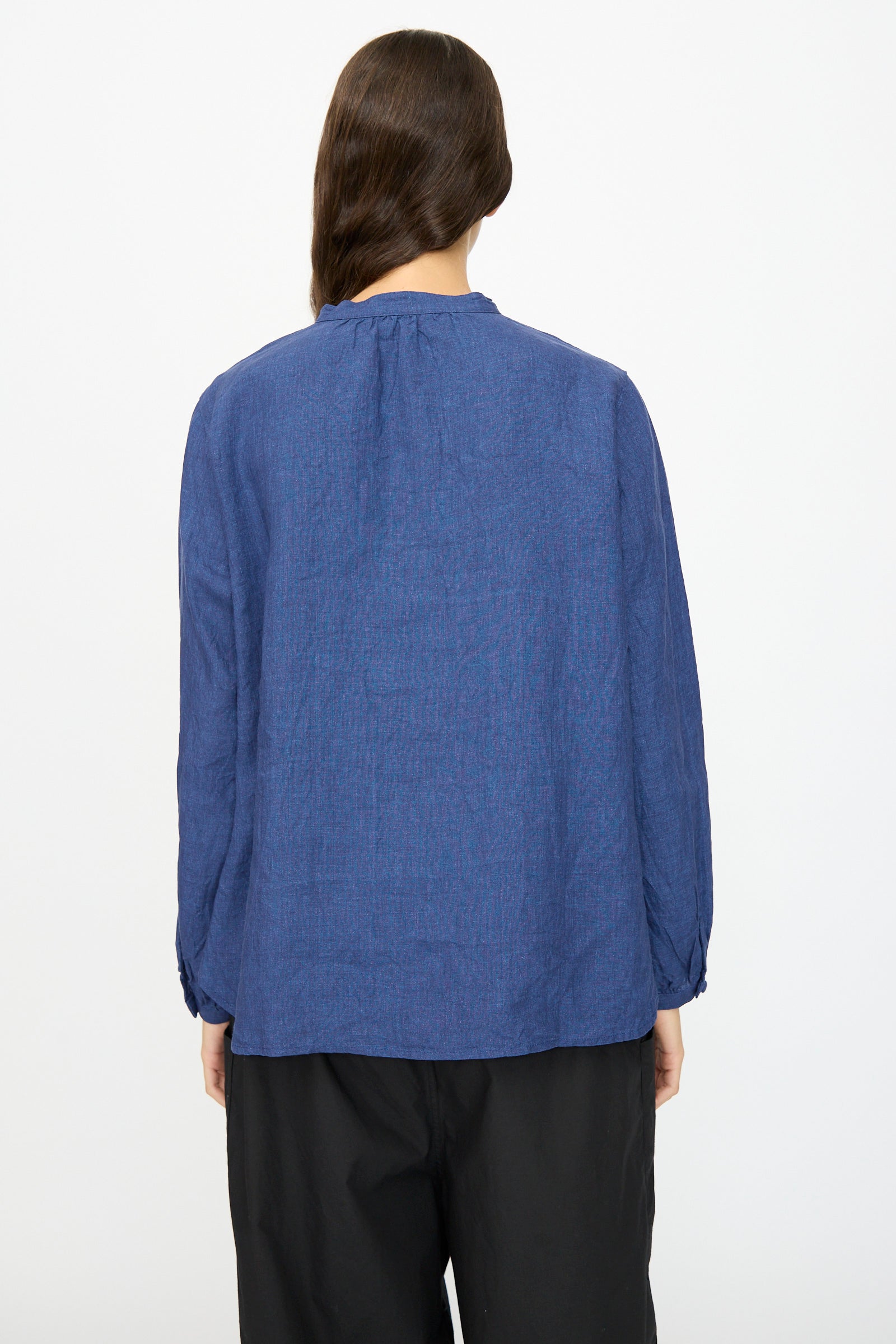 A person faces away from the camera against a white background, wearing Ichi Antiquités’ Linen Shirt in Indigo with a band collar, paired with black pants.