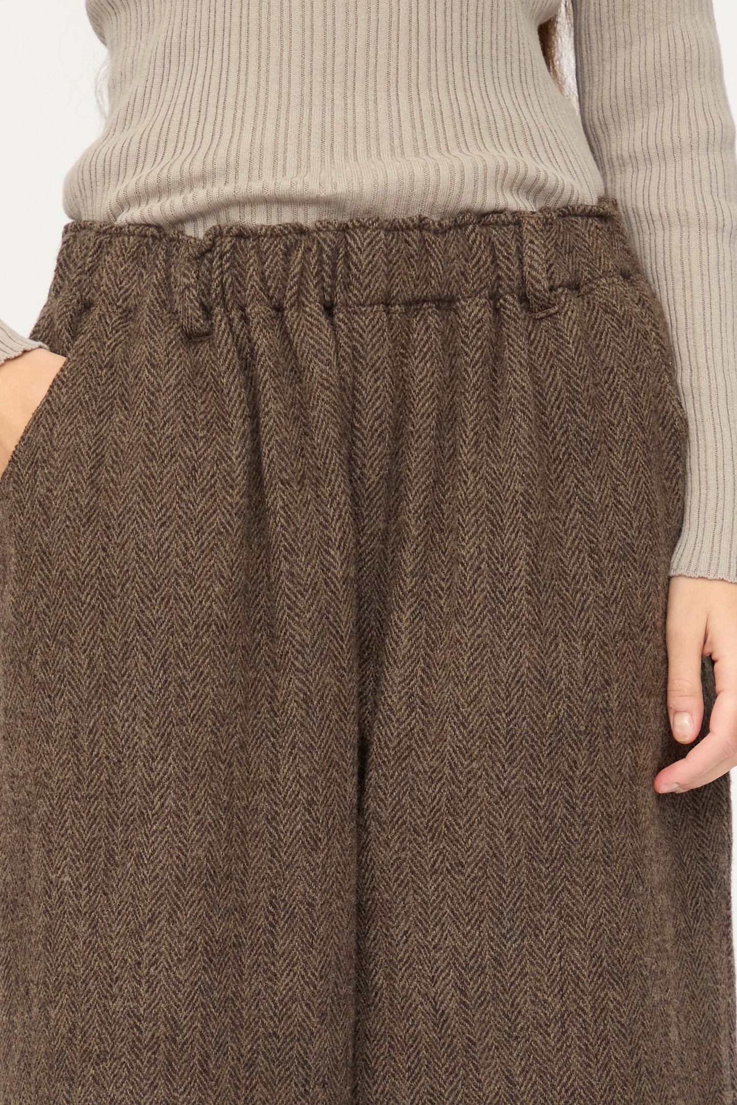 A person wears Ichi Antiquités Wool Herringbone Pants in Brown, featuring a high-waisted fit and pockets, paired with a light beige ribbed sweater, shown from mid-torso to mid-thigh.