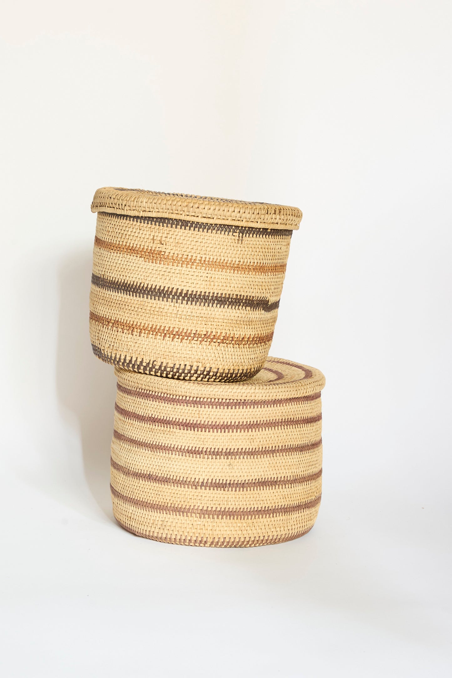 Two Motorohina Baskets with Yanomami stripe painting by Incausa, featuring horizontal brown and beige patterns, are stacked atop one another against a plain white background. Handwoven from natural materials, these Incausa baskets bring rustic elegance to any space.