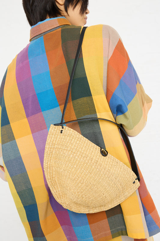 Person wearing a multicolored plaid coat and carrying an Inès Bressand N. 33 Medium Walnut Bag in Natural, a crescent-shaped, handwoven shoulder bag made from elephant grass.