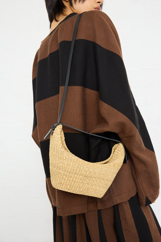 Woman carrying a N. 35 Mini Croissant Bag in Natural, handwoven from elephant grass, over her shoulder, dressed in a brown and black striped cape by Inès Bressand.
