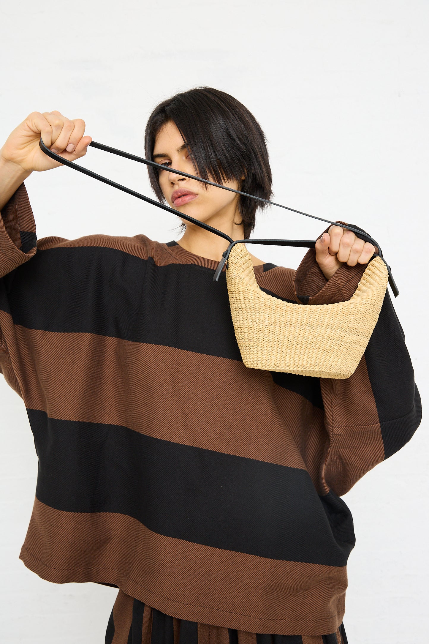 A woman pulling at the strap of a "N. 35 Mini Croissant Bag in Natural" handwoven bag slung over her shoulder by Inès Bressand.