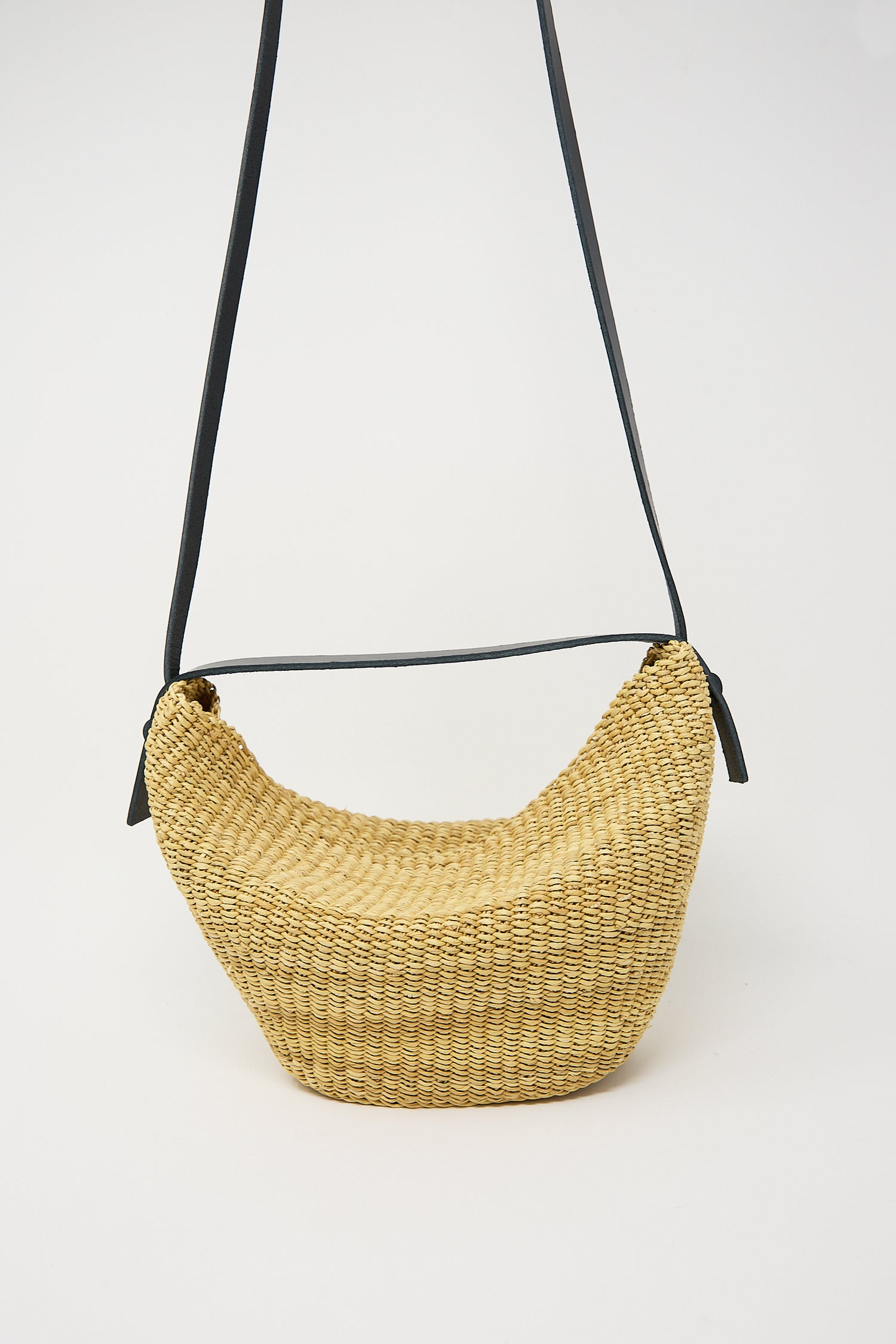 A handwoven straw N. 35 Mini Croissant Bag in Natural suspended from a thin black stand against a white background by Inès Bressand.