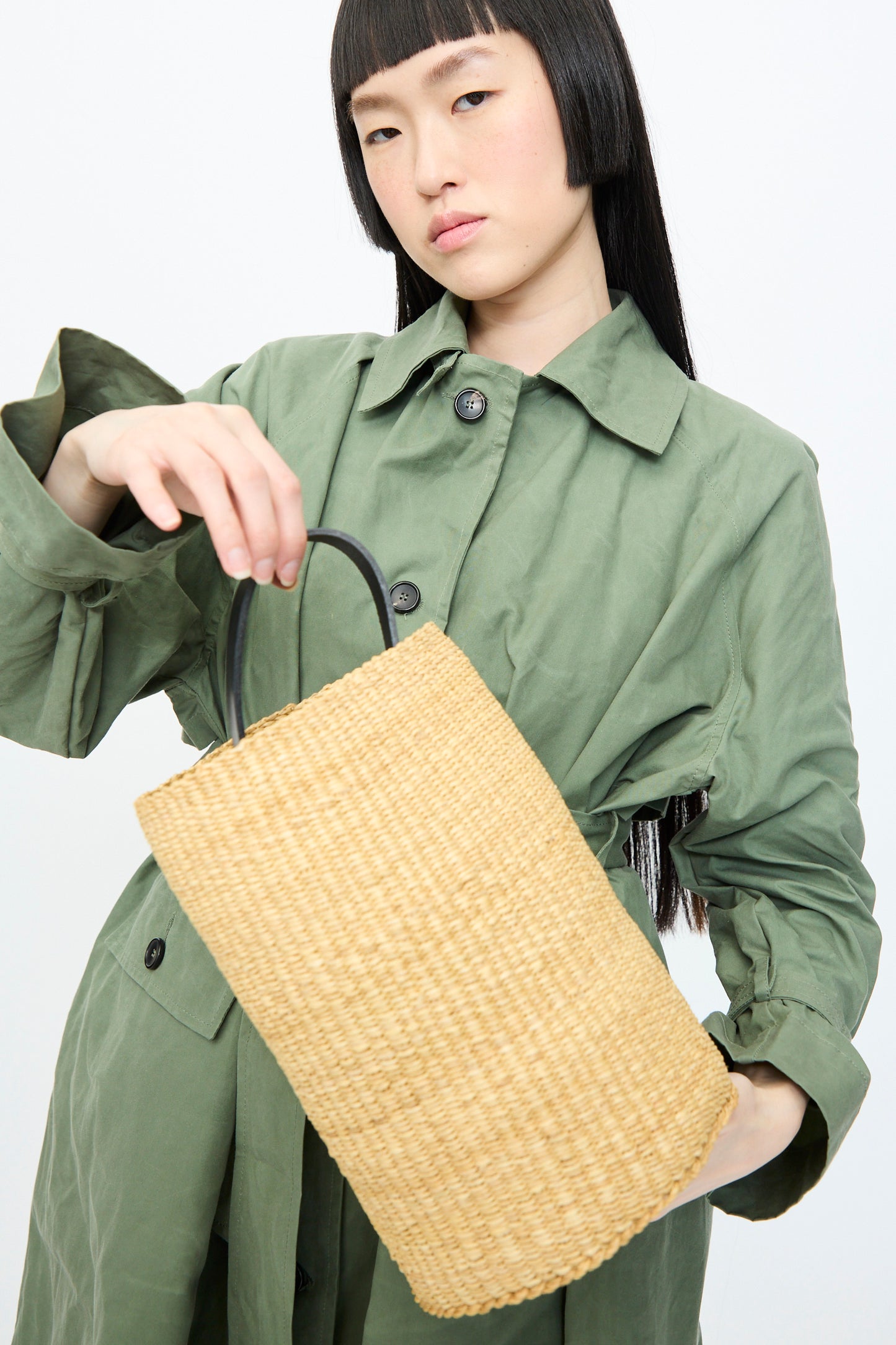 A person in a green coat holds the Seau Bag by Inès Bressand, made from handwoven elephant grass with black handles, against a plain background.