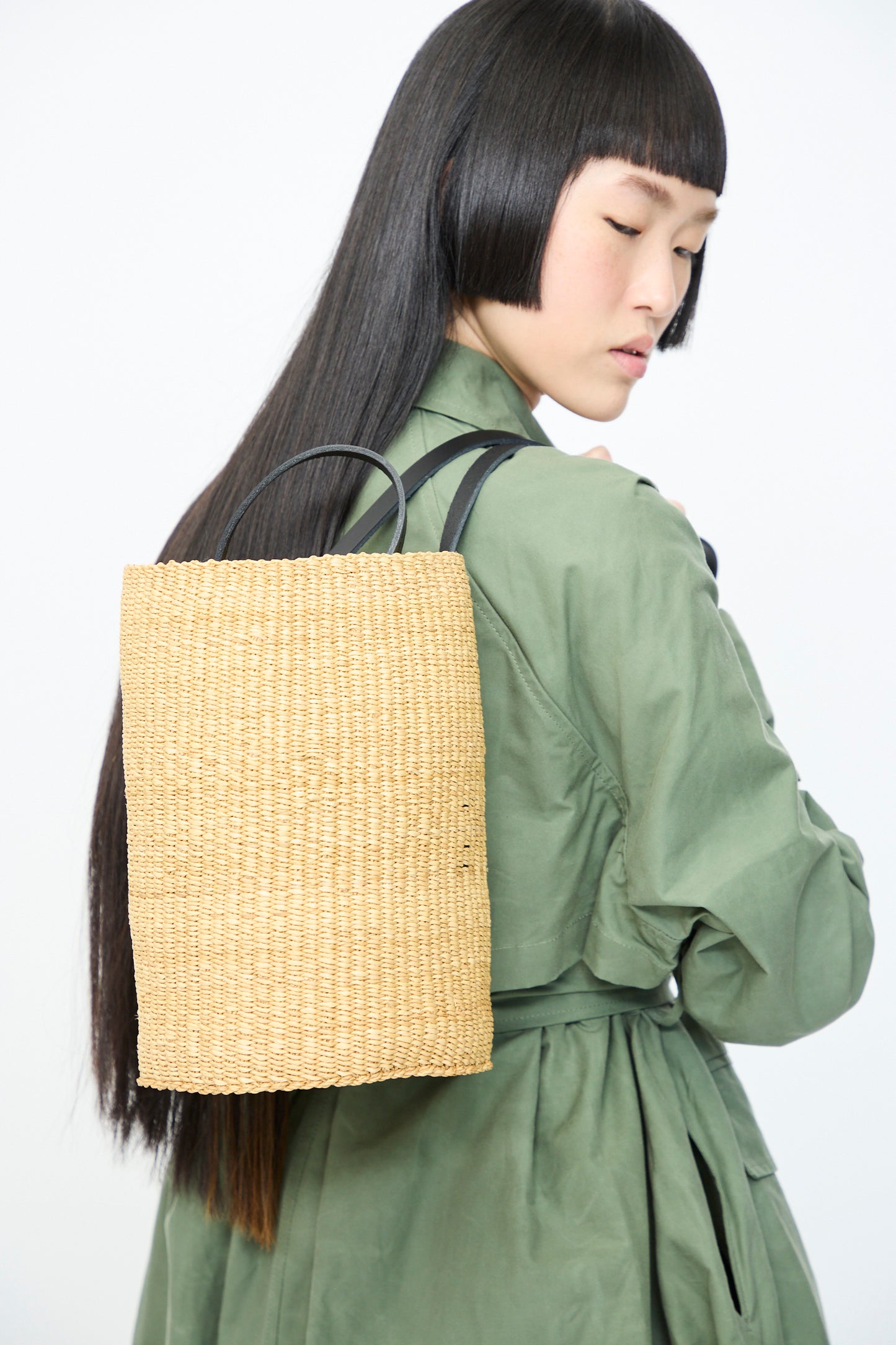 A person with long black hair in a green coat carries the Seau Bag in Natural with Black Natural, made from handwoven elephant grass by Inès Bressand. They glance back over their shoulder against a plain backdrop.