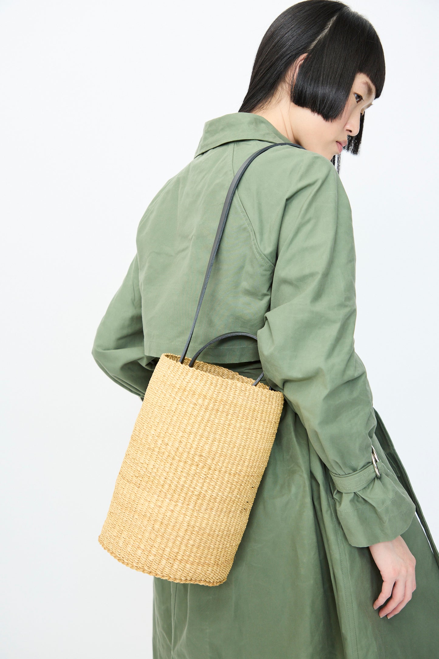 A person in a green trench coat with a short bob hairstyle carries the Inès Bressand Seau Bag in Natural with Black Natural over their shoulder, glancing to the side against a plain background.