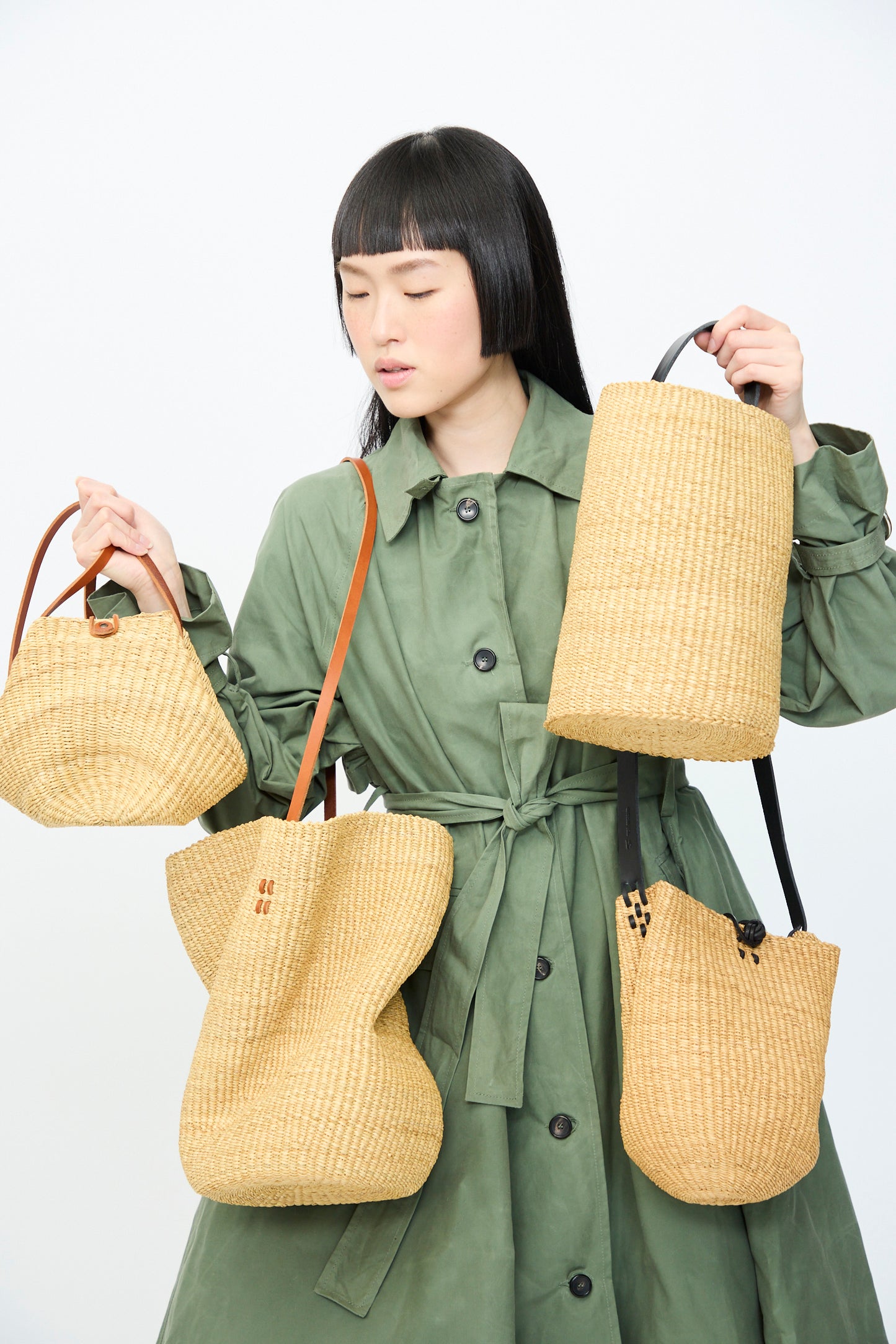 A person in a green trench coat stands against a plain backdrop, highlighting four straw bags with brown straps. One is the Seau Bag in Natural with Black Natural by Inès Bressand.