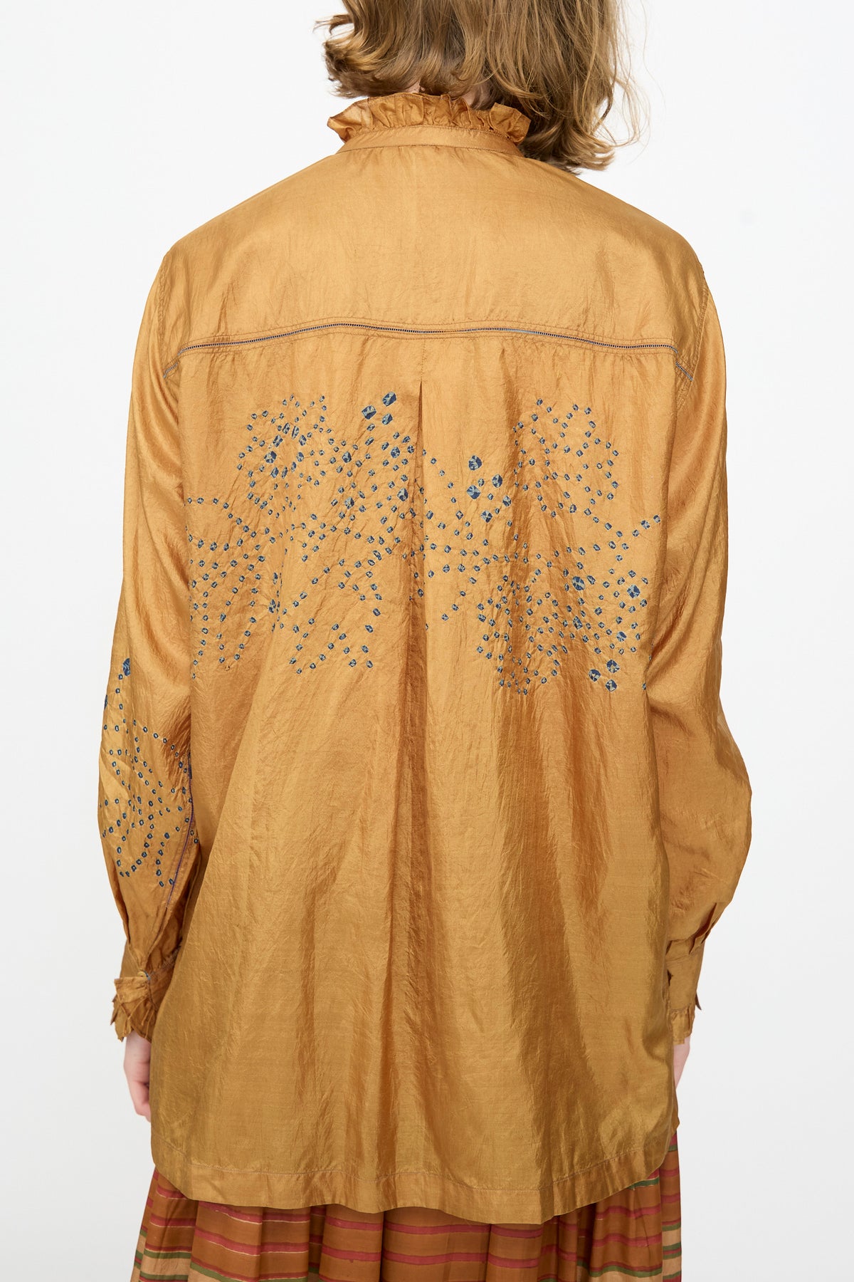 A person wearing the Injiri Bandhani Silk Henri Shirt in Ochre—a loose-fitting, handcrafted silk shirt with a shiny golden-brown hue, ruffled collar and cuffs, and small blue floral embroidery adorning the back and sleeves.