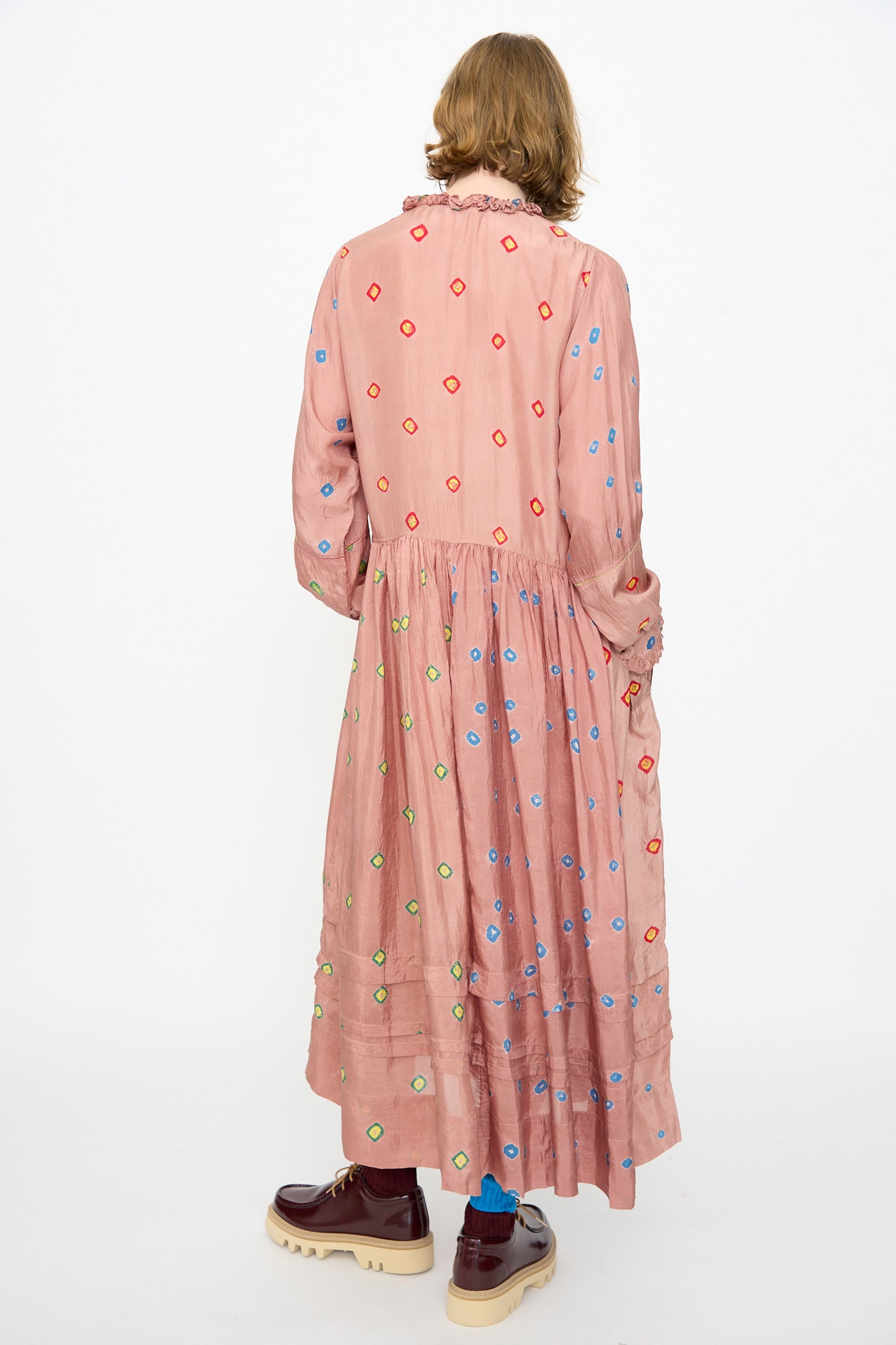 A person wearing the Injiri Bandhani Silk Natalie Dress in Mauve, a handcrafted maxi dress featuring colorful embroidered dots, paired with brown platform shoes and blue socks, stands with their back to the camera against a plain white background.