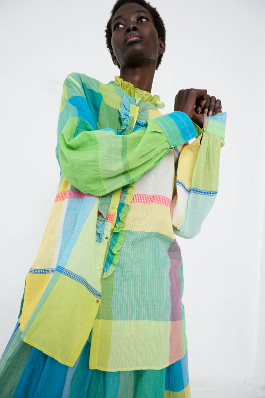 A woman in an Injiri cotton shirt in green, yellow, and pink check stands against a white background, gazing thoughtfully to the side.