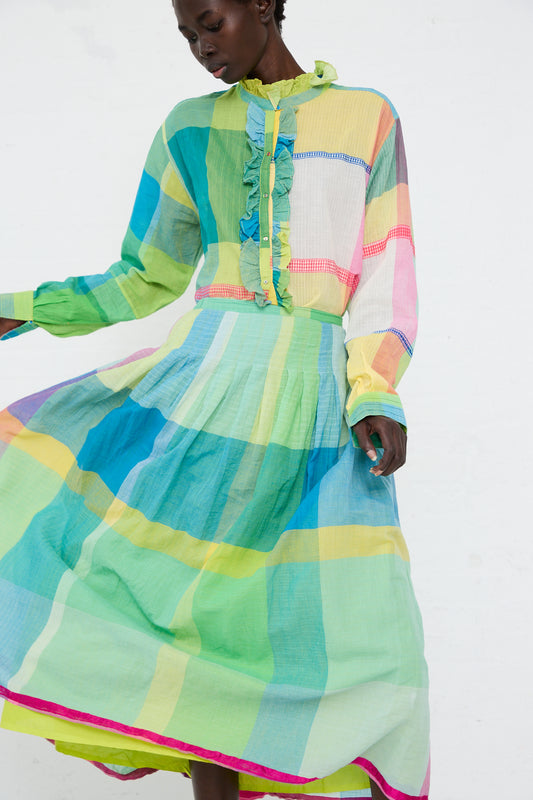 A woman in a vibrant Injiri cotton skirt in green, blue and yellow check with a hood, looking downwards, against a plain white background.