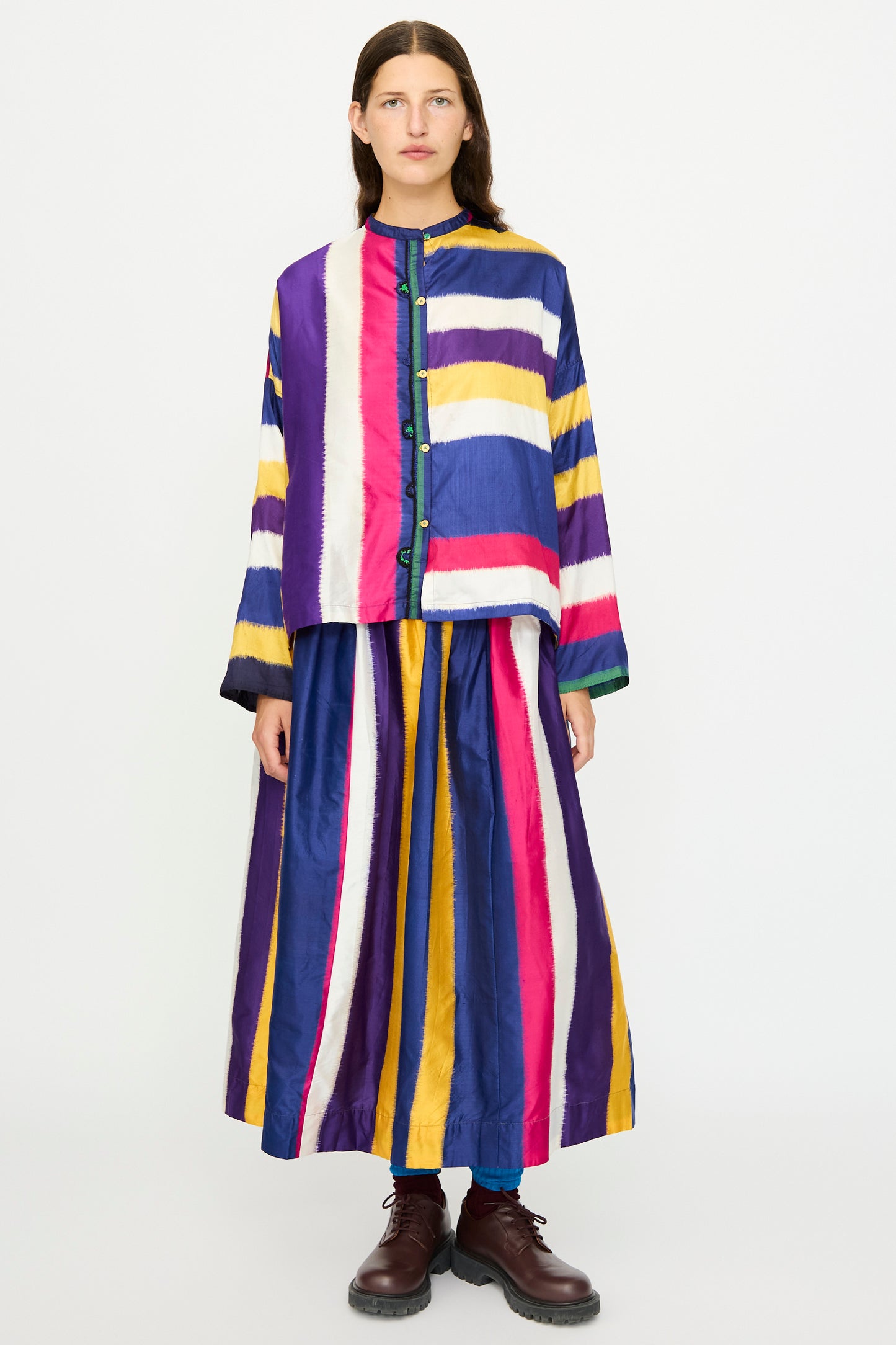 A woman faces forward wearing the Injiri Ikat Silk Shirt in Multi Stripe, a boxy fit silk design with a band collar and bold vertical and horizontal stripes in blue, yellow, purple, white, and magenta.