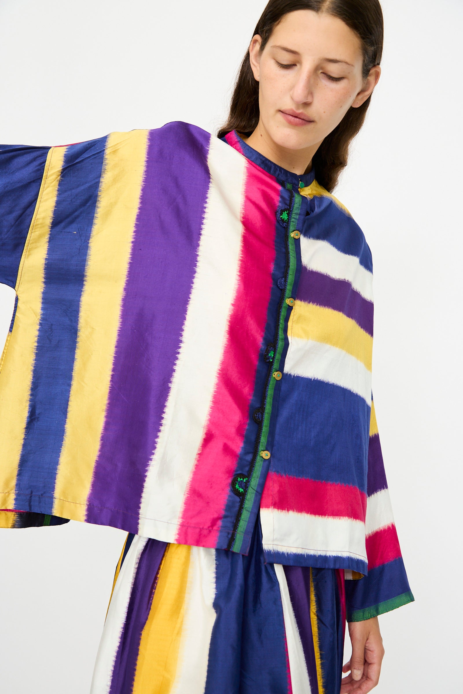 A person wears the Injiri Ikat Silk Shirt in Multi Stripe, featuring loose long sleeves and bold vertical stripes in yellow, purple, white, blue, and red, standing against a plain background.