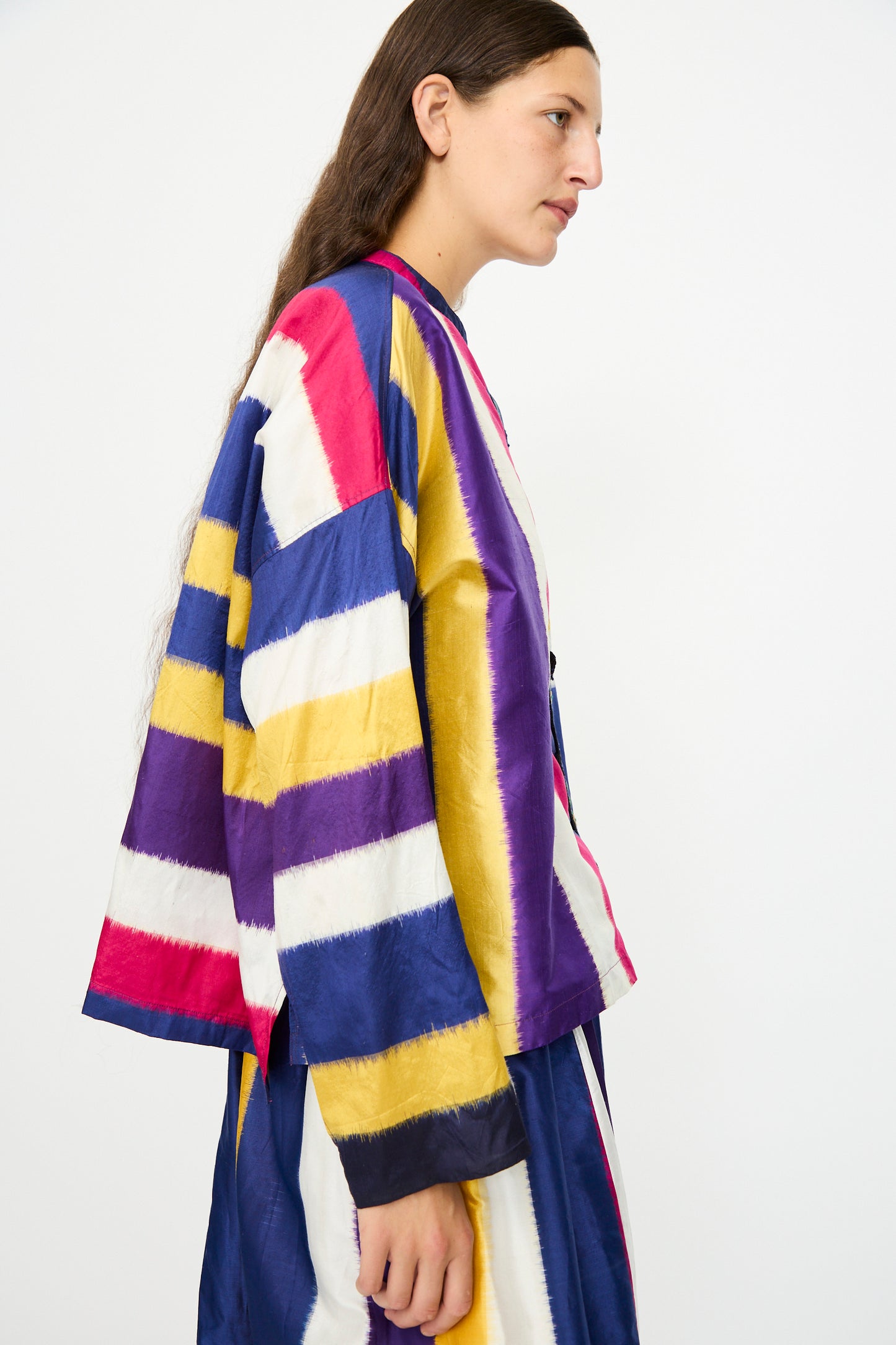 A person with long brown hair wears the Injiri Ikat Silk Shirt in Multi Stripe, featuring a boxy fit and wide horizontal stripes, paired with a loose jacket, standing against a plain white background.