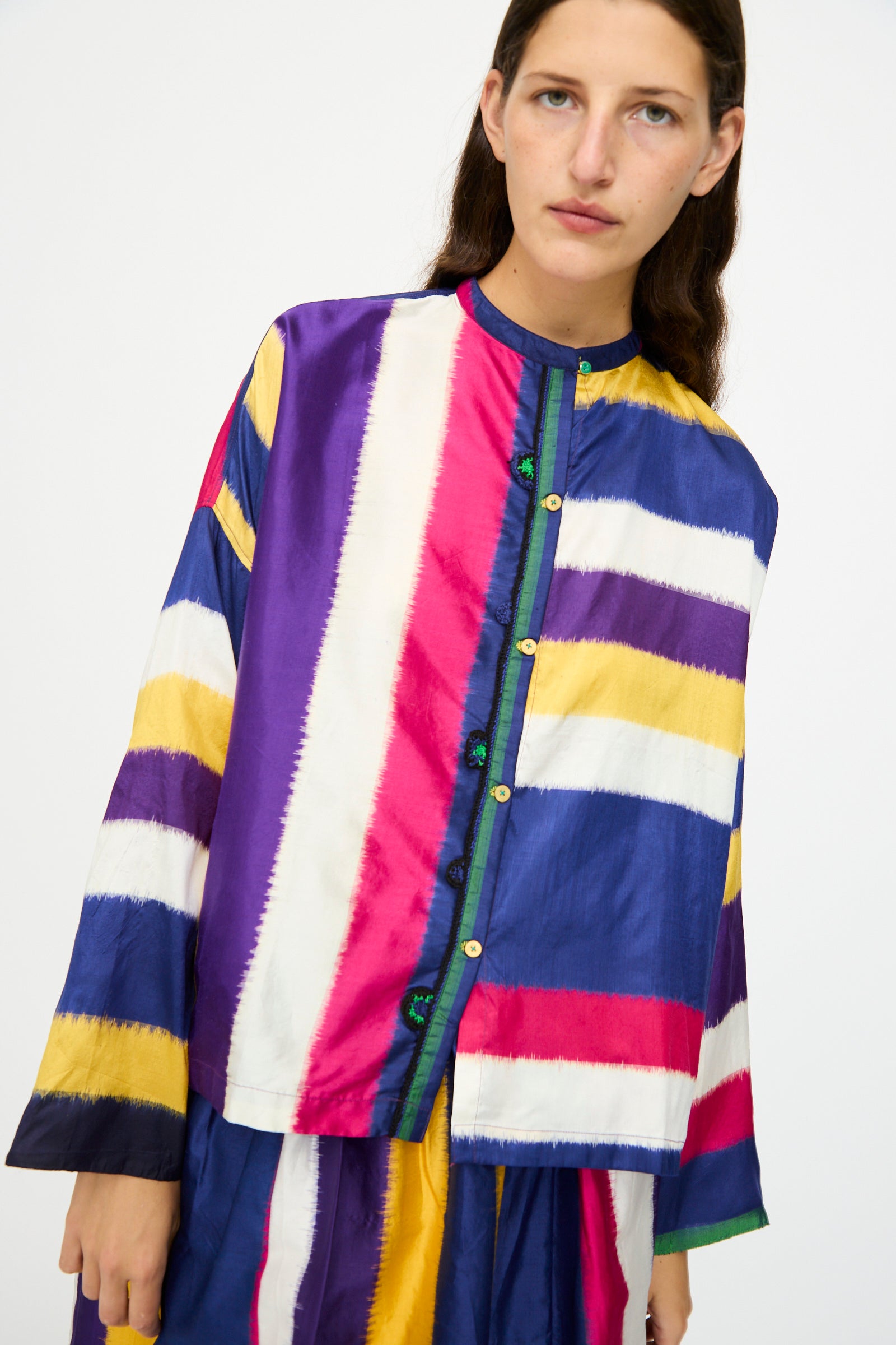 A person wears the Injiri Ikat Silk Shirt in Multi Stripe, featuring long sleeves and blue, purple, yellow, red, and white patterns, standing against a plain background.