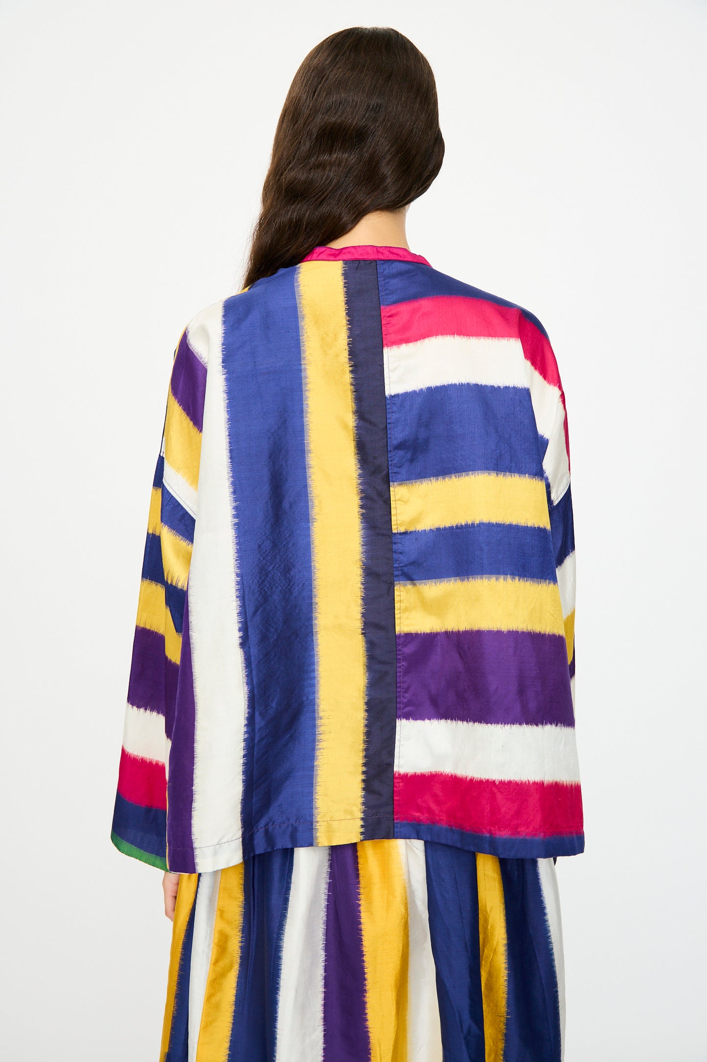A woman with long brown hair, seen from behind, wears the Injiri Ikat Silk Shirt in Multi Stripe—boxy fit with bold horizontal and vertical stripes in yellow, blue, purple, white, and red.
