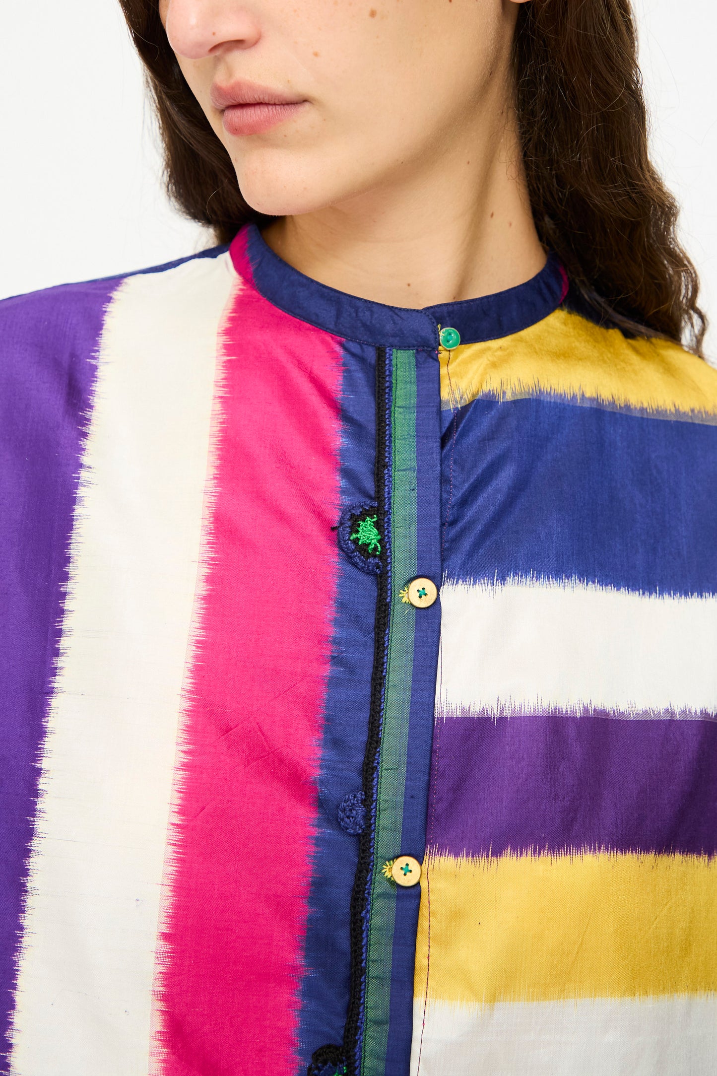 A woman wears the Injiri Ikat Silk Shirt in Multi Stripe, featuring vertical stripes in purple, white, pink, yellow, and blue. This boxy-fit silk shirt highlights contrasting buttons and unique collar detailing.