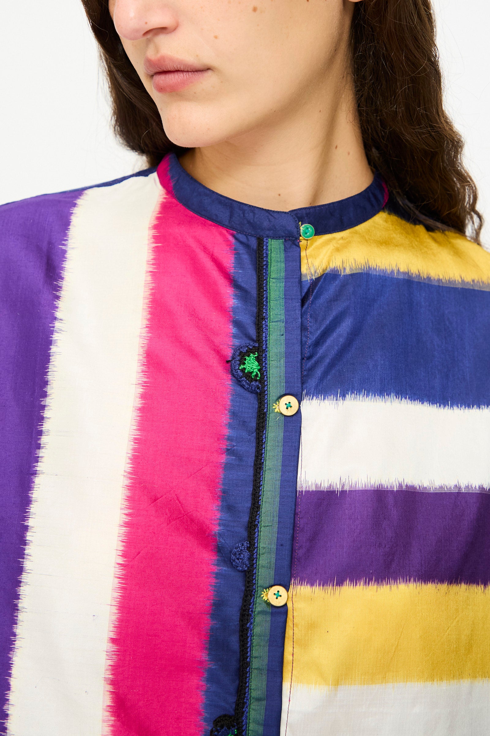 A woman wears the Injiri Ikat Silk Shirt in Multi Stripe, featuring vertical stripes in purple, white, pink, yellow, and blue. This boxy-fit silk shirt highlights contrasting buttons and unique collar detailing.