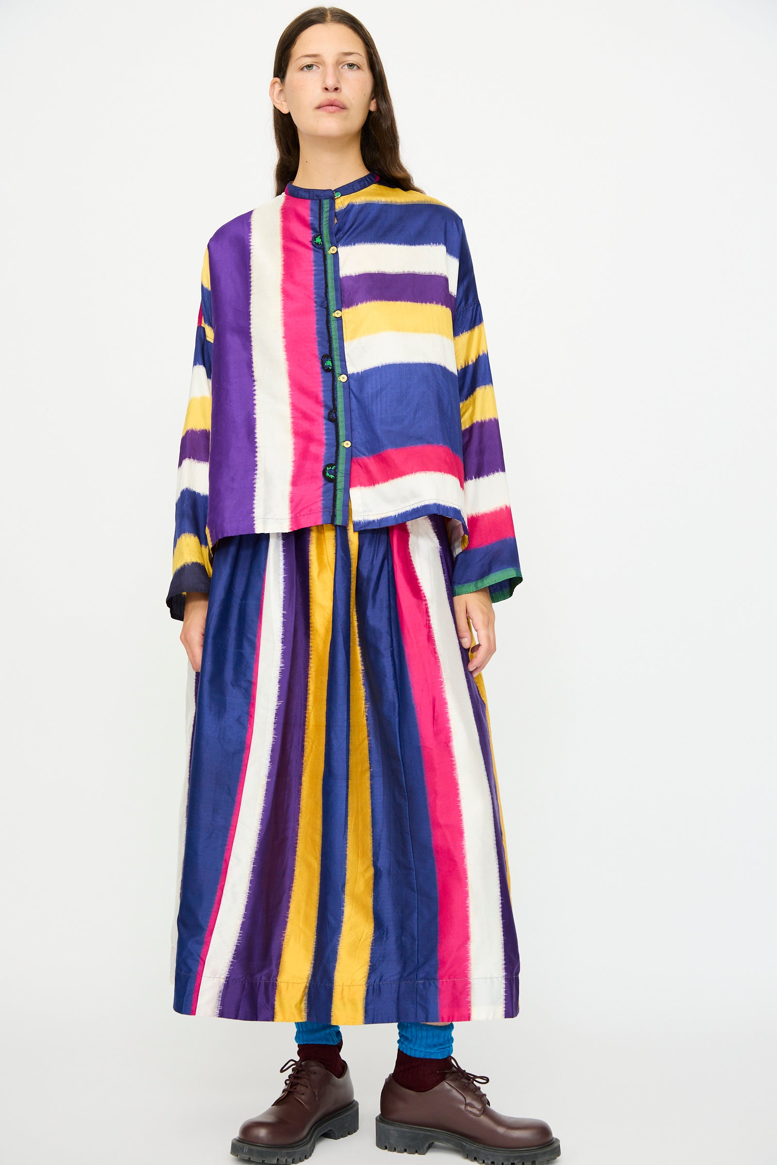 A person stands against a plain white background, wearing Injiri's Ikat Silk Skirt lined with Cotton in Multi Stripe, paired with a button-up top, dark brown shoes, and blue socks.