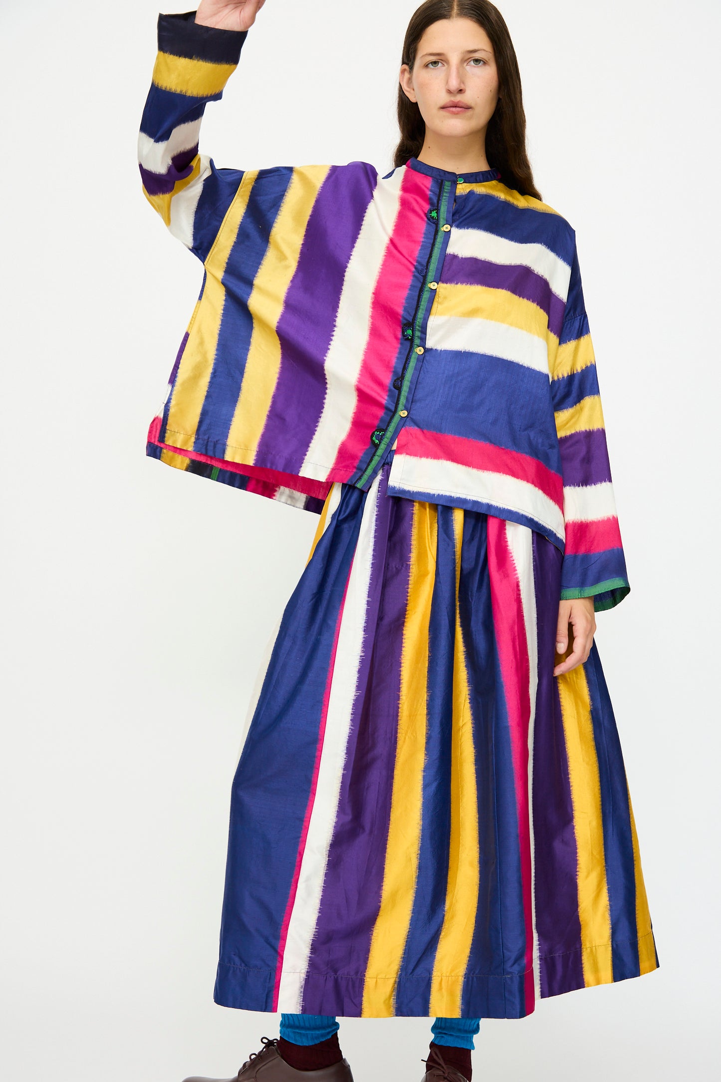 A person wears the Injiri Ikat Silk Skirt lined with cotton in multi stripe, styled with wide sleeves and an A-line silhouette, standing against a plain white background.