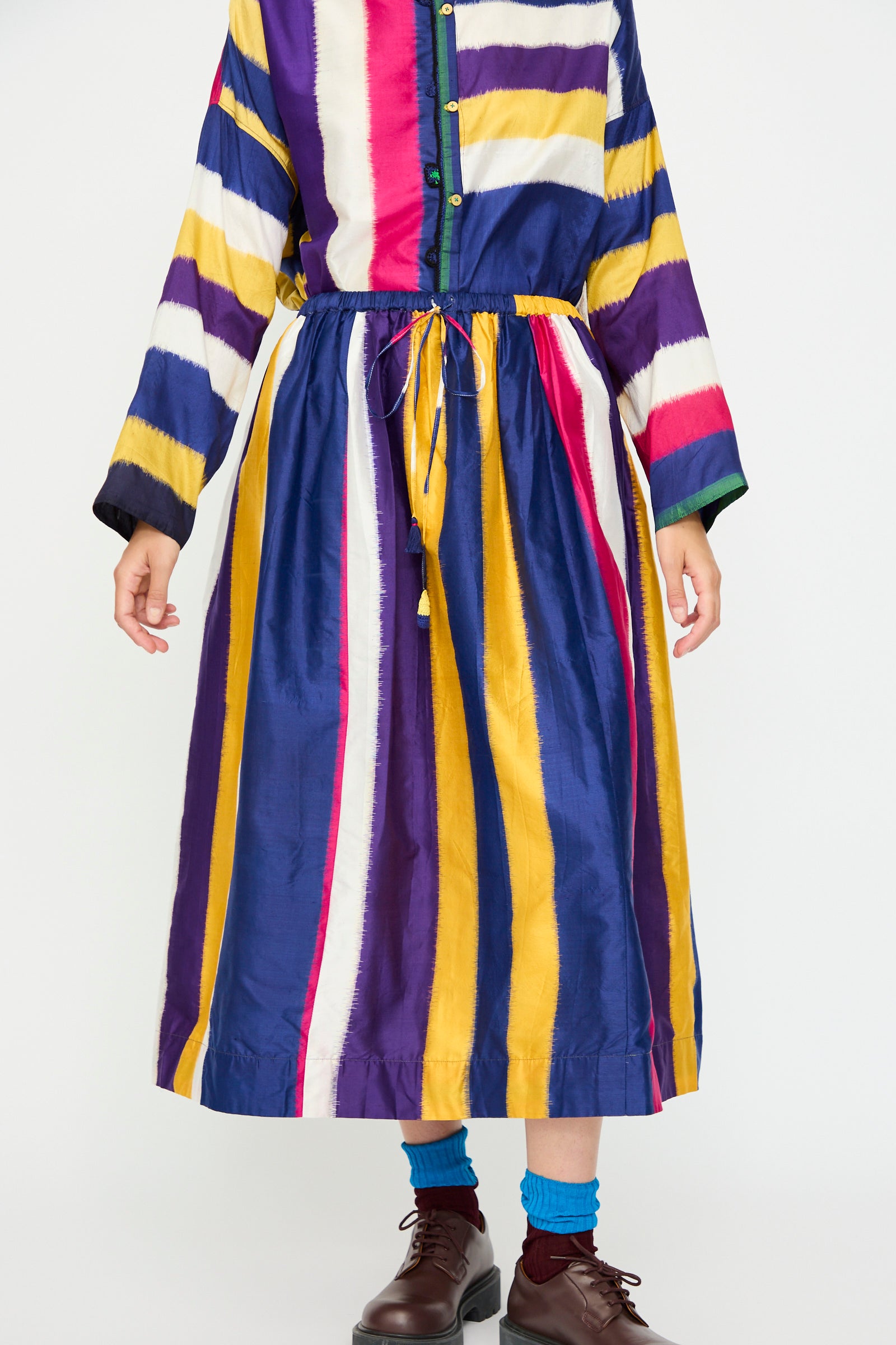 Person wearing the Injiri Ikat Silk Skirt lined with Cotton in Multi Stripe, featuring vertical yellow, blue, pink, and white stripes. The look is paired with blue socks and brown shoes; photo cropped at the shoulders.