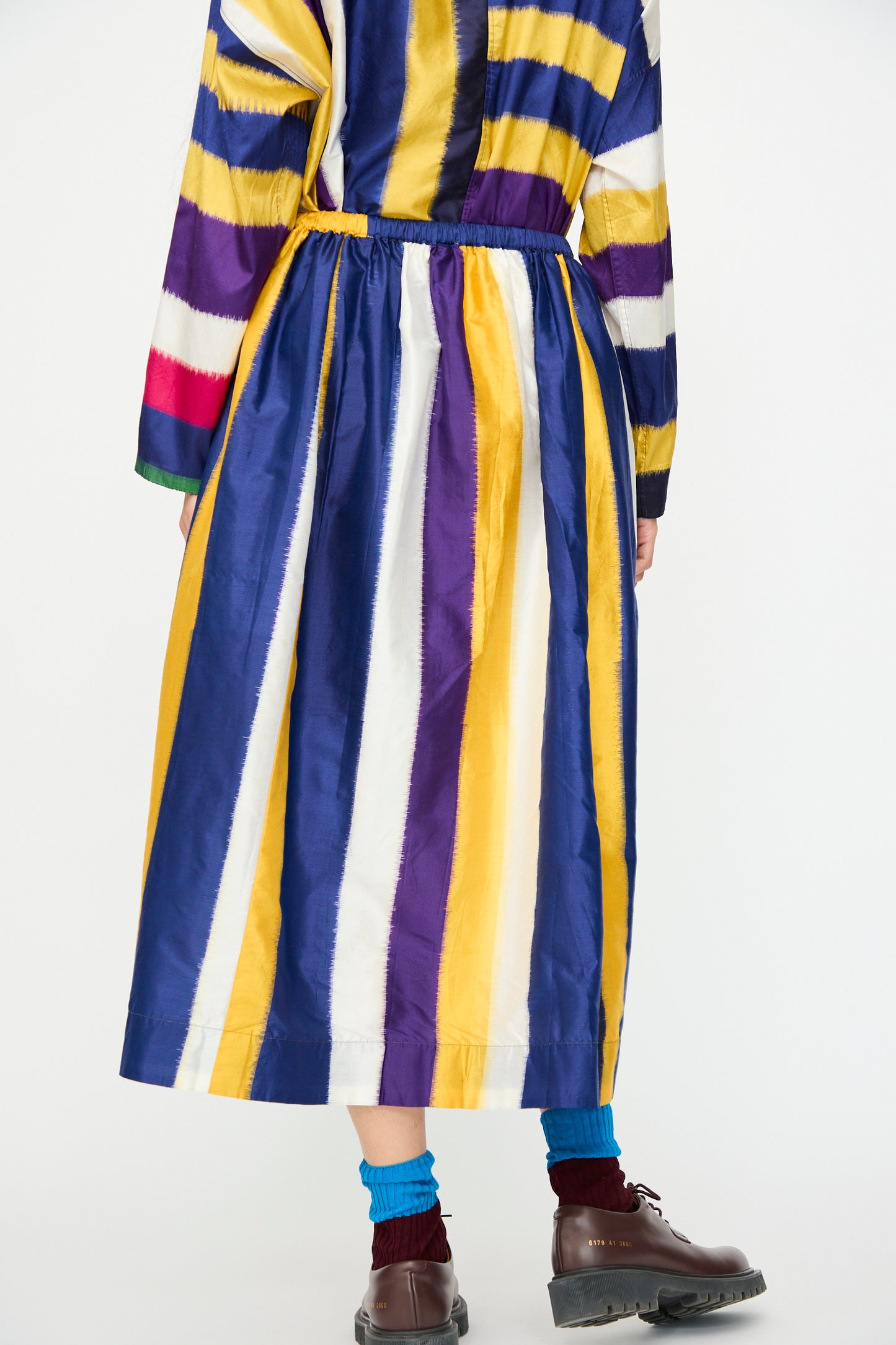 A person wears the Injiri Ikat Silk Skirt lined with Cotton in Multi Stripe, featuring blue, yellow, white, and purple stripes, paired with brown shoes and teal socks, standing against a white background.