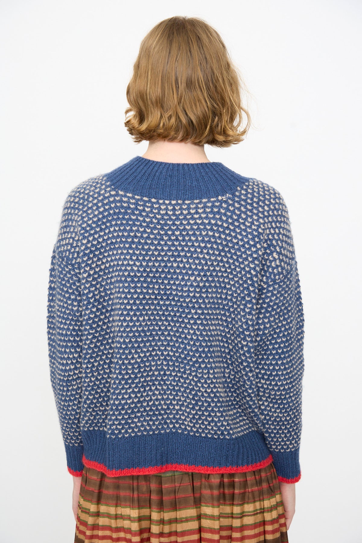 A person with short, light brown hair is shown from behind, wearing the Injiri Knitted Wool Sweater in Blue with white knit details and a brown striped skirt against a plain white background.
