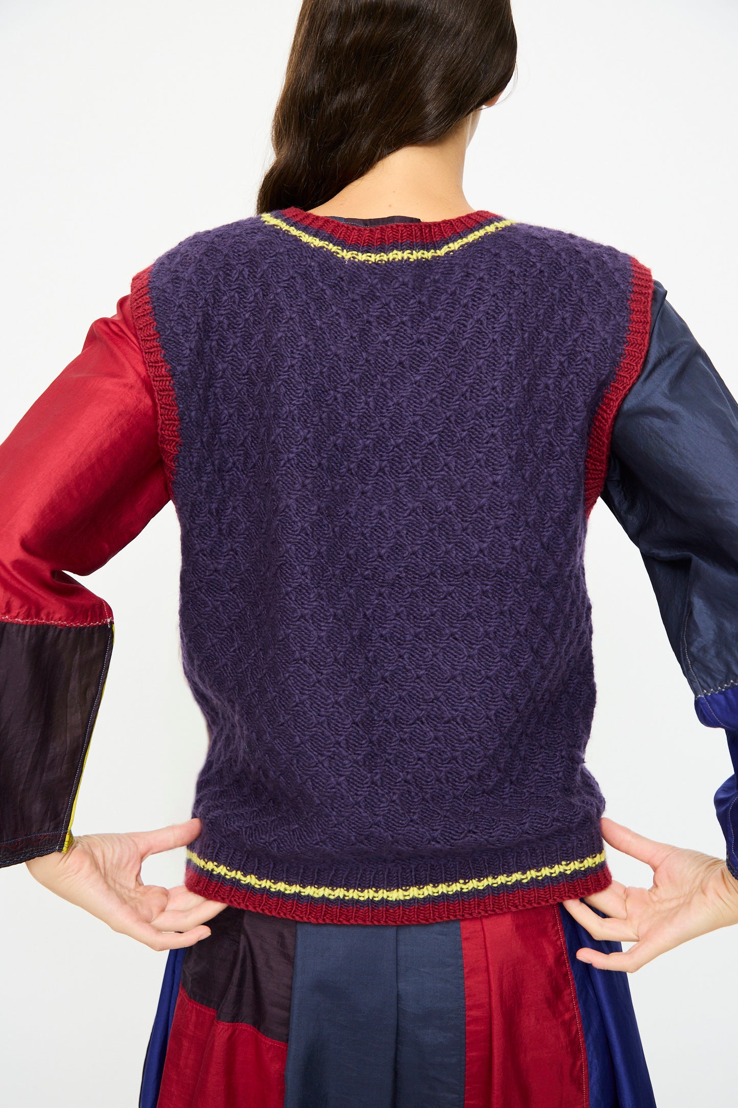 A person faces away, wearing the Injiri Merino Wool Sweater Vest in Purple, Red and Yellow over a long-sleeve multicolored dress.