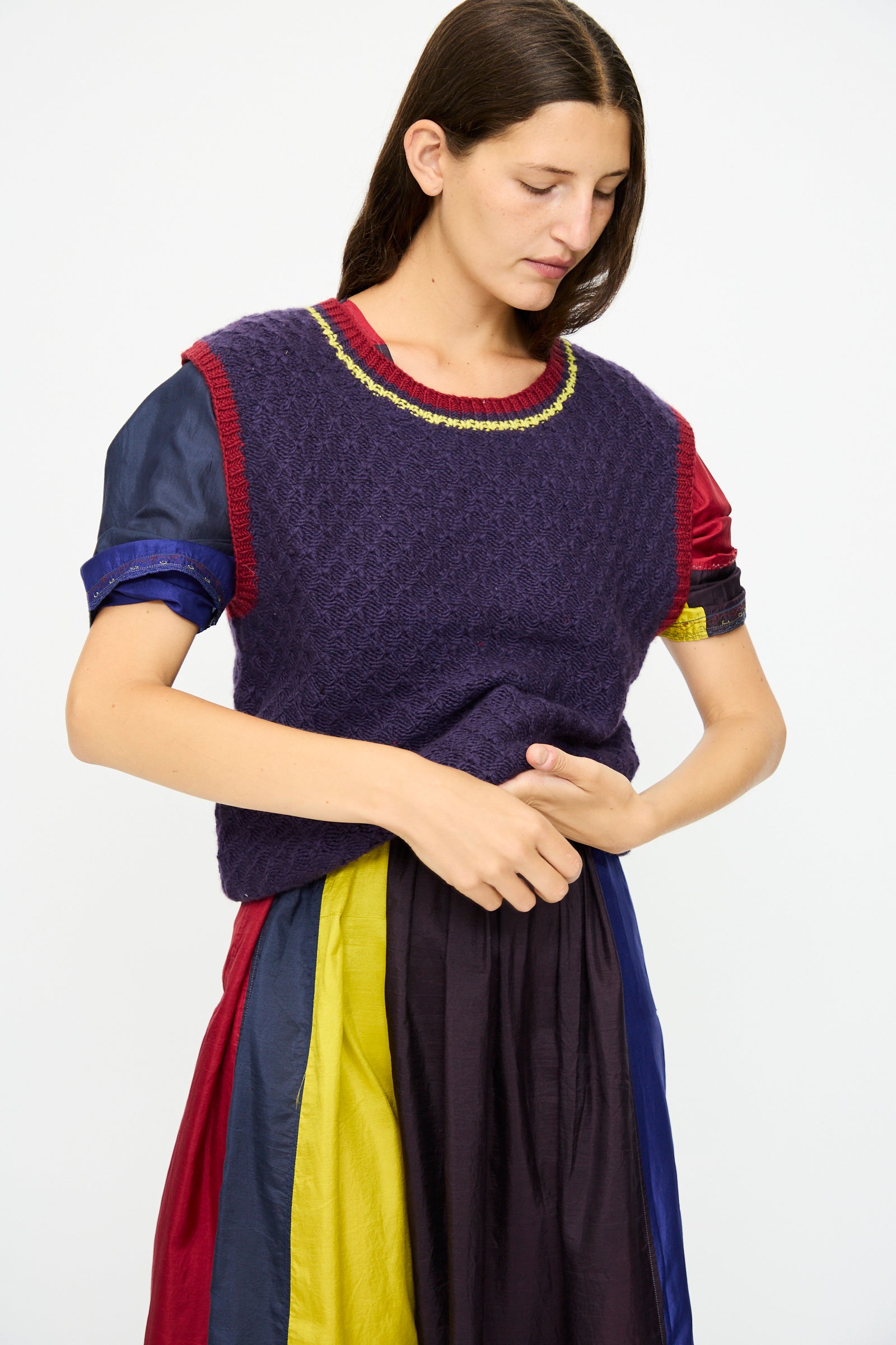 A person with long brown hair wears the Injiri Merino Wool Sweater Vest in Purple, Red and Yellow over a multicolored, short-sleeve dress while standing against a plain white background.