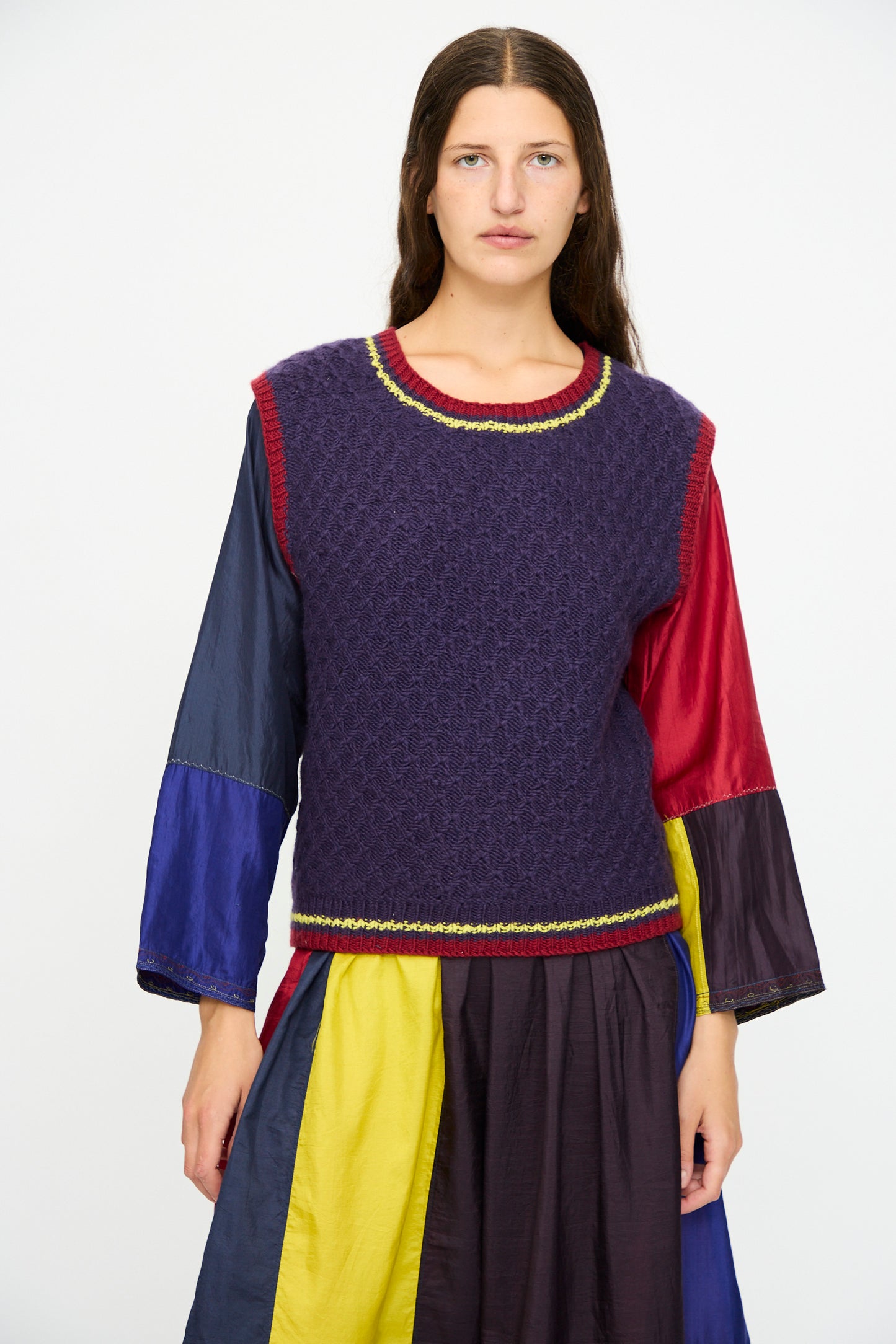 A woman stands against a plain background wearing the Injiri Merino Wool Sweater Vest in purple, red, and yellow, layered over a multicolored dress with long sleeves and a relaxed fit.