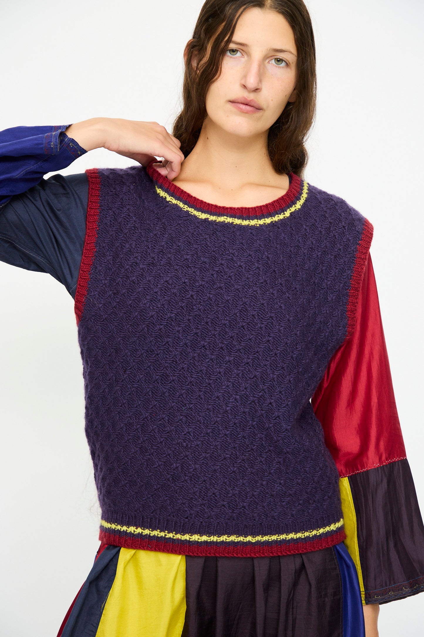 A woman with long brown hair wears the Injiri Merino Wool Sweater Vest in purple, red, and yellow over a multicolored long-sleeve dress.